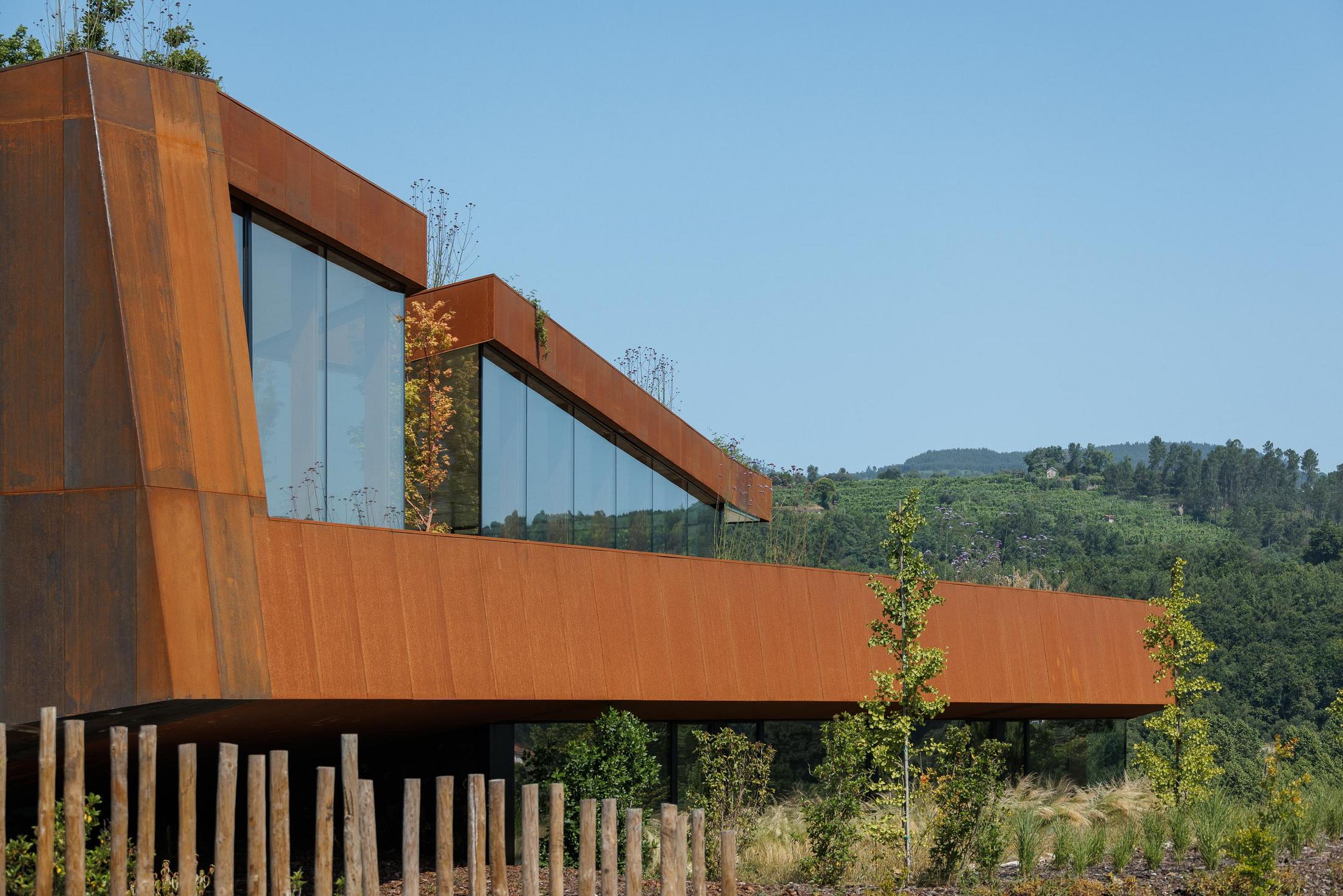 Exterior: Fachada da casa com revestimento em aço corten, grandes janelas e estrutura suspensa sobre pilares. — Casa Corten arquitetura HPA Arquitetura e Investimentos em Celorico de Basto