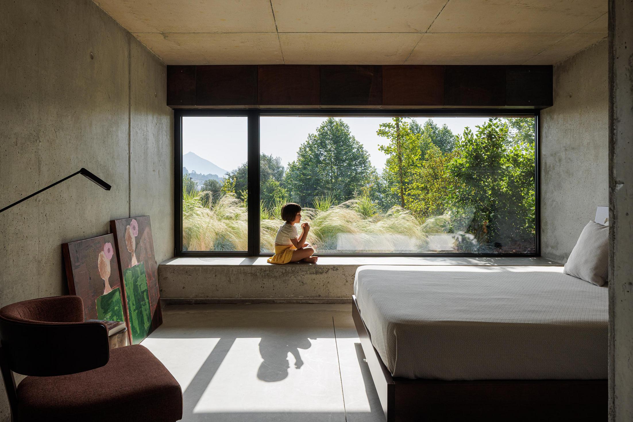 Quarto: Interior minimalista com paredes em betão aparente, cama de casal e grande janela com vista para o exterior. — Casa Corten arquitetura HPA Arquitetura e Investimentos em Celorico de Basto
