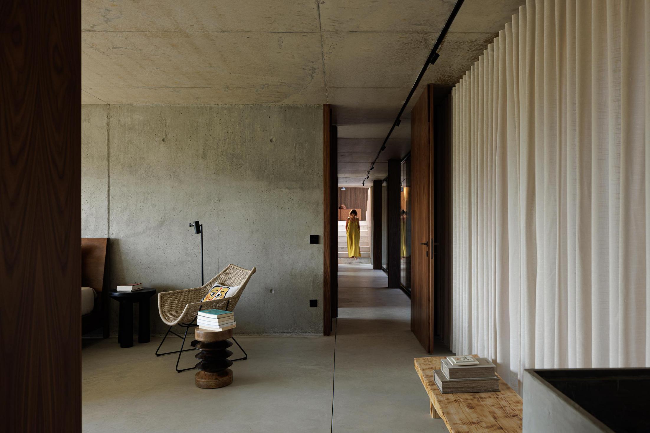 Quarto: Espaço amplo com cama, cadeirão de vime, e acesso a corredor com cortinas brancas e paredes em betão. — Casa Corten arquitetura HPA Arquitetura e Investimentos em Celorico de Basto