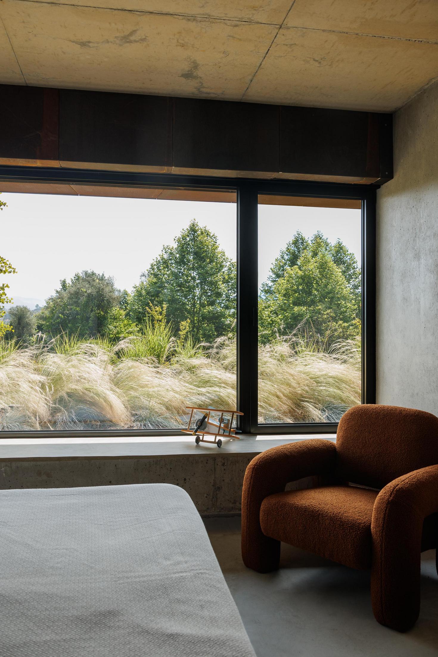 Quarto: Detalhe de janela com vista para o exterior, cadeirão castanho e cama com roupa de cama branca. — Casa Corten arquitetura HPA Arquitetura e Investimentos em Celorico de Basto
