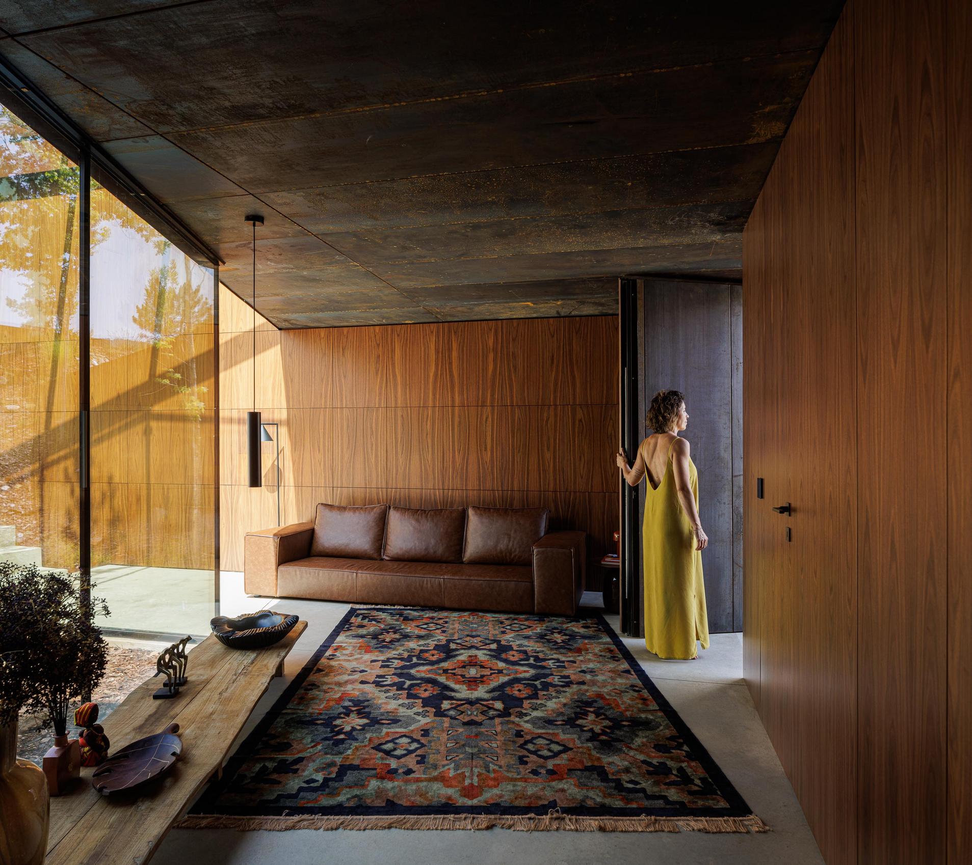 Sala de estar: Interior com sofá castanho, alcatifa colorida, paredes em madeira e grande janela com vista para o exterior. — Casa Corten arquitetura HPA Arquitetura e Investimentos em Celorico de Basto