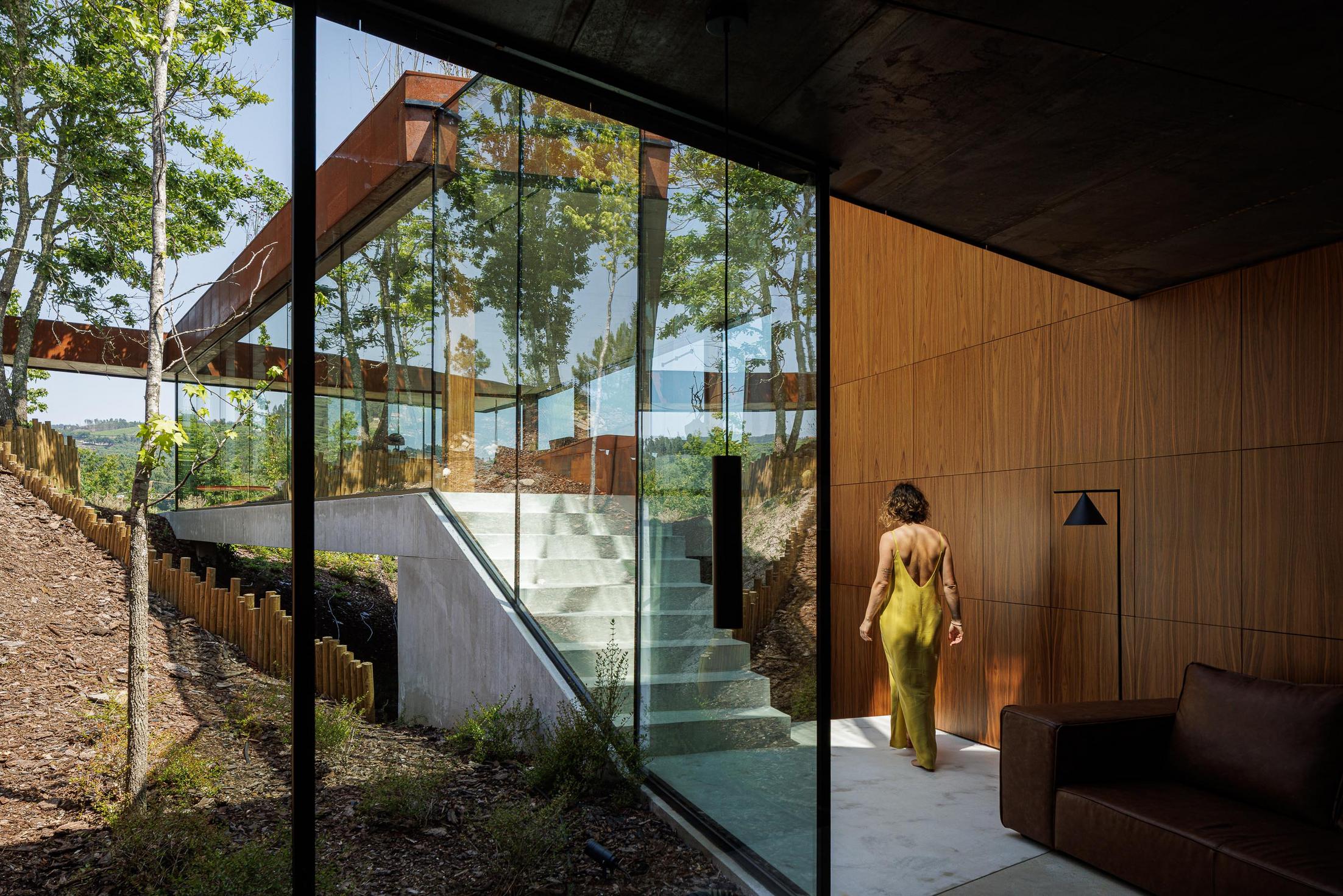 Sala de estar: Interior com sofá castanho, paredes em madeira e acesso a escadas exteriores com vista para a paisagem. — Casa Corten arquitetura HPA Arquitetura e Investimentos em Celorico de Basto