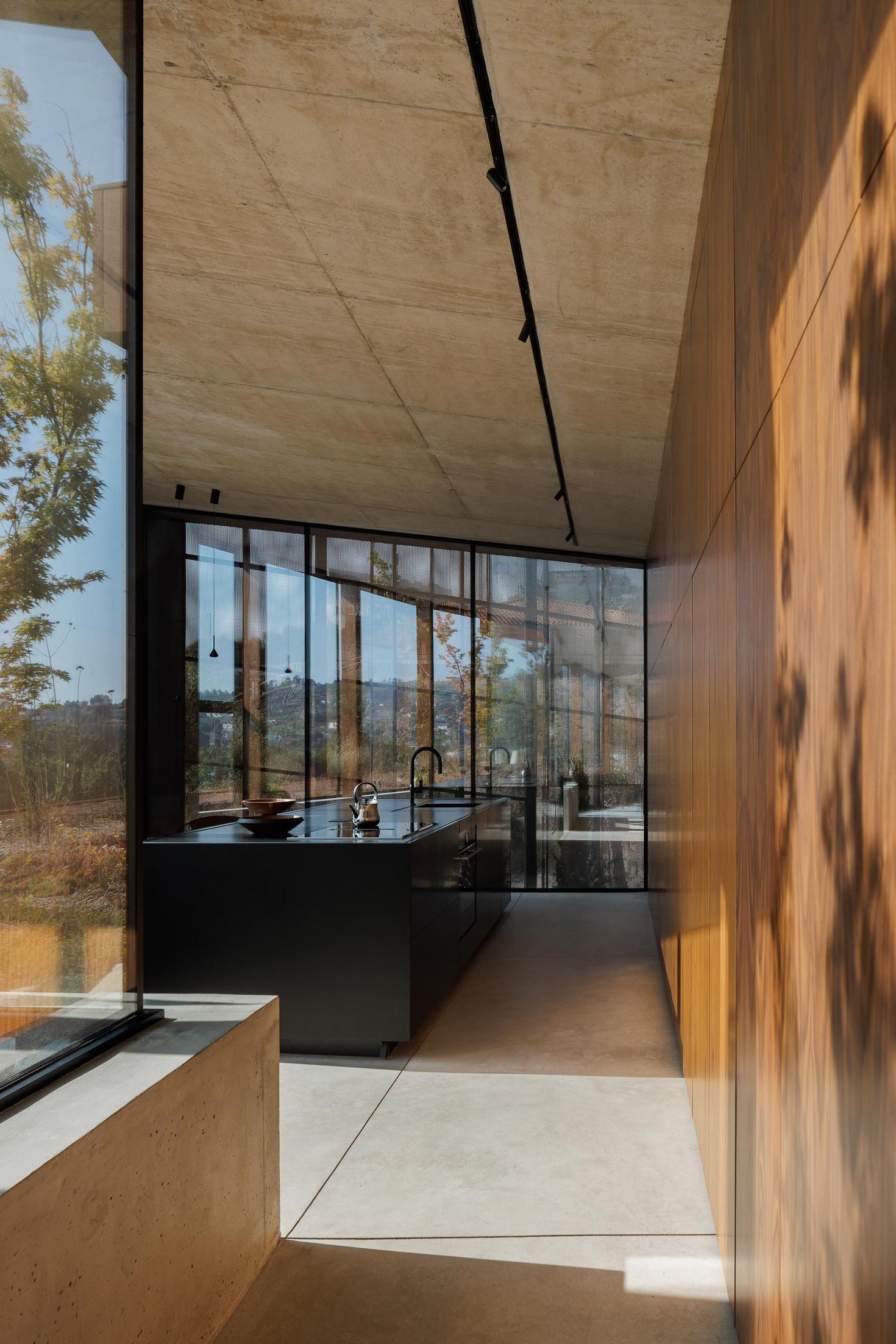 Cozinha: Interior com ilha central preta, paredes em madeira e grande janela com vista para o exterior. — Casa Corten arquitetura HPA Arquitetura e Investimentos em Celorico de Basto