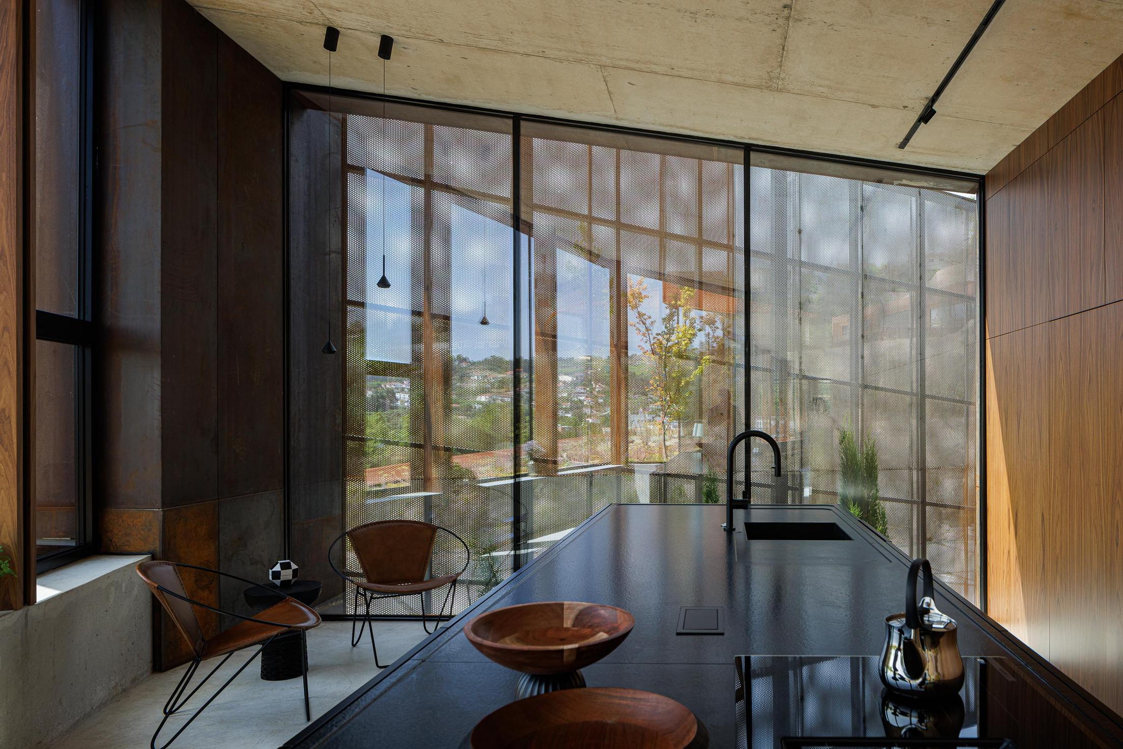 Cozinha: Ilha central preta com lavatório, cadeiras de design e janela com tela metálica perfurada. — Casa Corten arquitetura HPA Arquitetura e Investimentos em Celorico de Basto