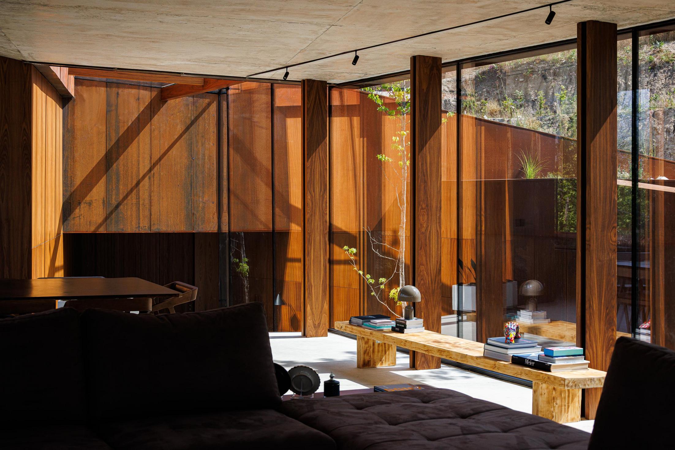 Sala de estar: Interior com sofá castanho, mesa de madeira, paredes em madeira e grande janela com vista para o exterior. — Casa Corten arquitetura HPA Arquitetura e Investimentos em Celorico de Basto