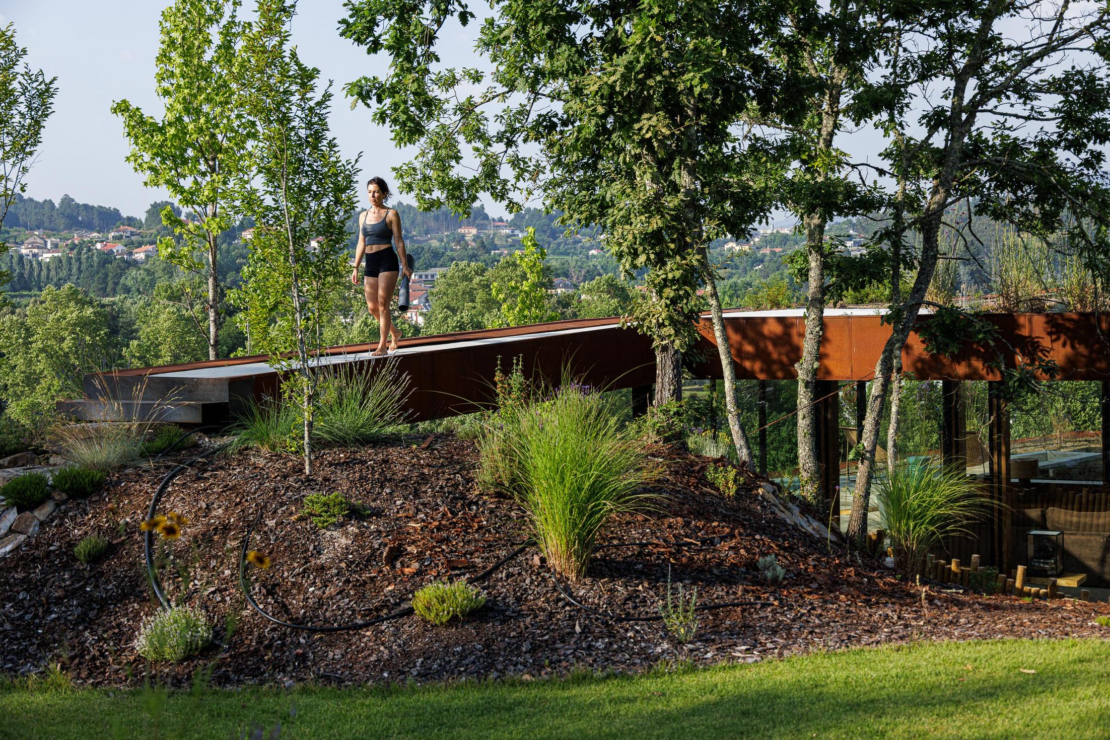 Exterior: Mulher a caminhar no telhado verde da casa, com vegetação rasteira e árvores. — Casa Corten arquitetura HPA Arquitetura e Investimentos em Celorico de Basto
