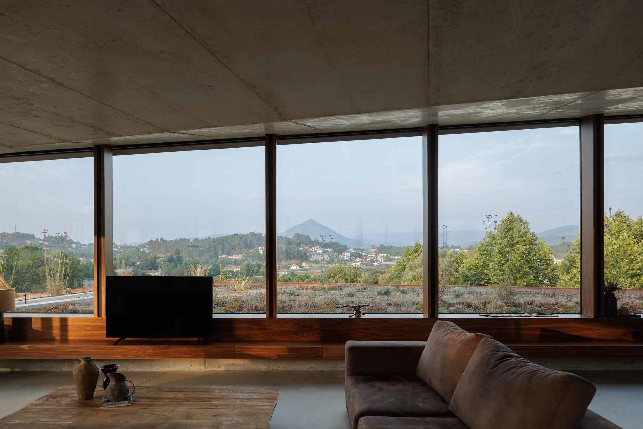Sala de estar: Interior com sofá castanho, mesa de madeira e grande janela com vista para a paisagem ao anoitecer. — Casa Corten arquitetura HPA Arquitetura e Investimentos em Celorico de Basto