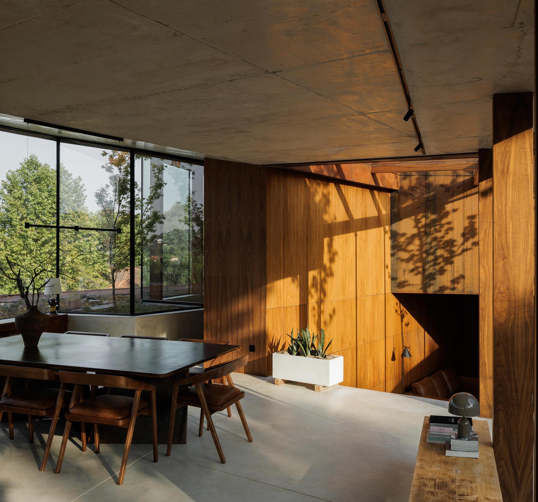 Sala de jantar: Interior com mesa de jantar em madeira, cadeiras de design e vista para o exterior ao anoitecer. — Casa Corten arquitetura HPA Arquitetura e Investimentos em Celorico de Basto
