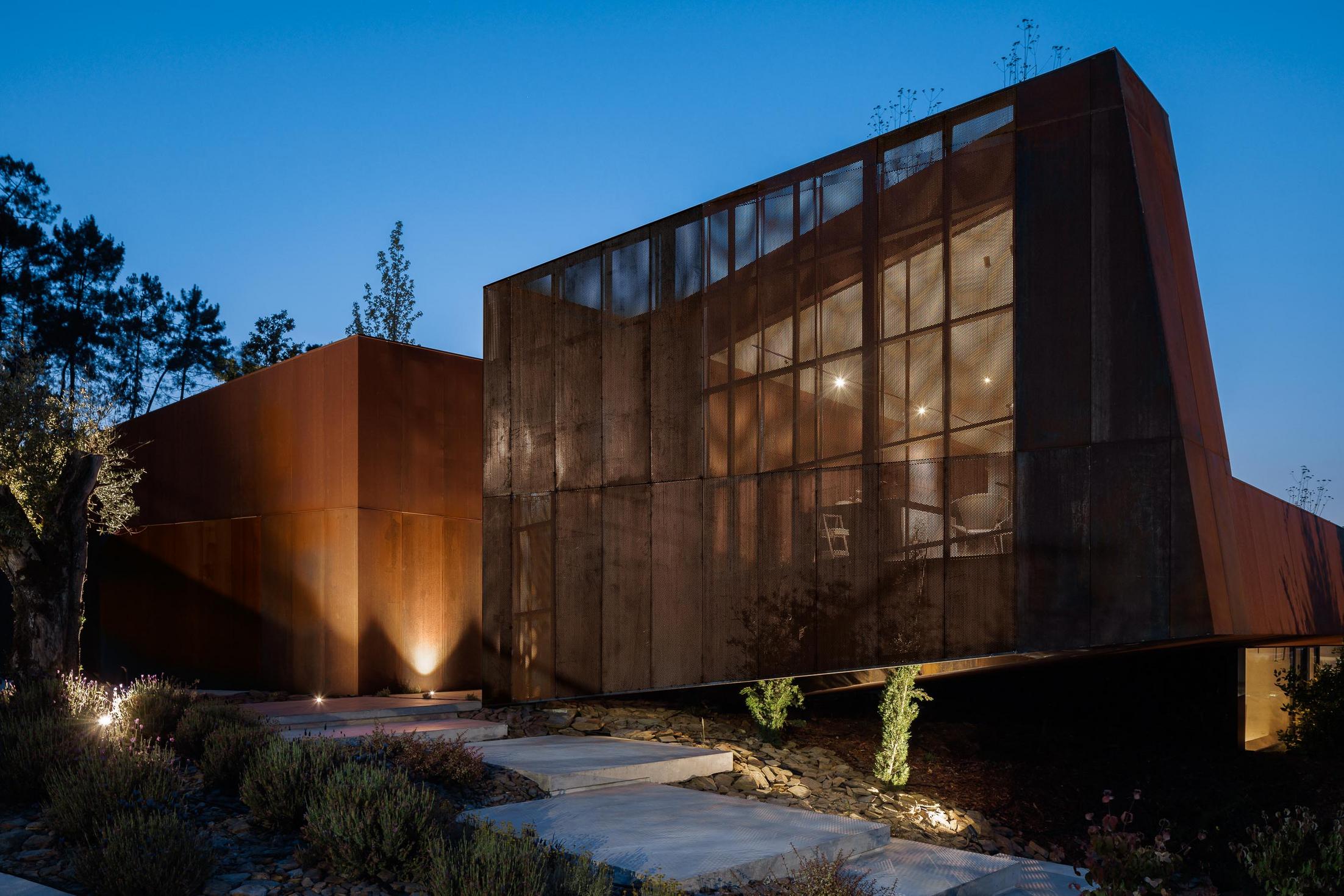 Exterior: Detalhe da fachada em aço corten, com vegetação rasteira e uma pequena árvore em primeiro plano. — Casa Corten arquitetura HPA Arquitetura e Investimentos em Celorico de Basto