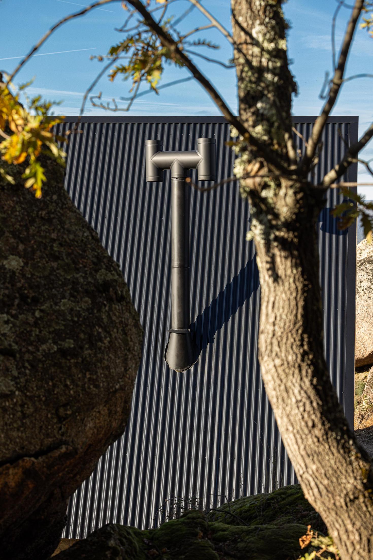 Exterior da cabana com chaminé preta e revestimento metálico, entre rochas e árvores. — Abrigos de Montanha arquitetura Filipe Pina Arquitectura em Belmonte