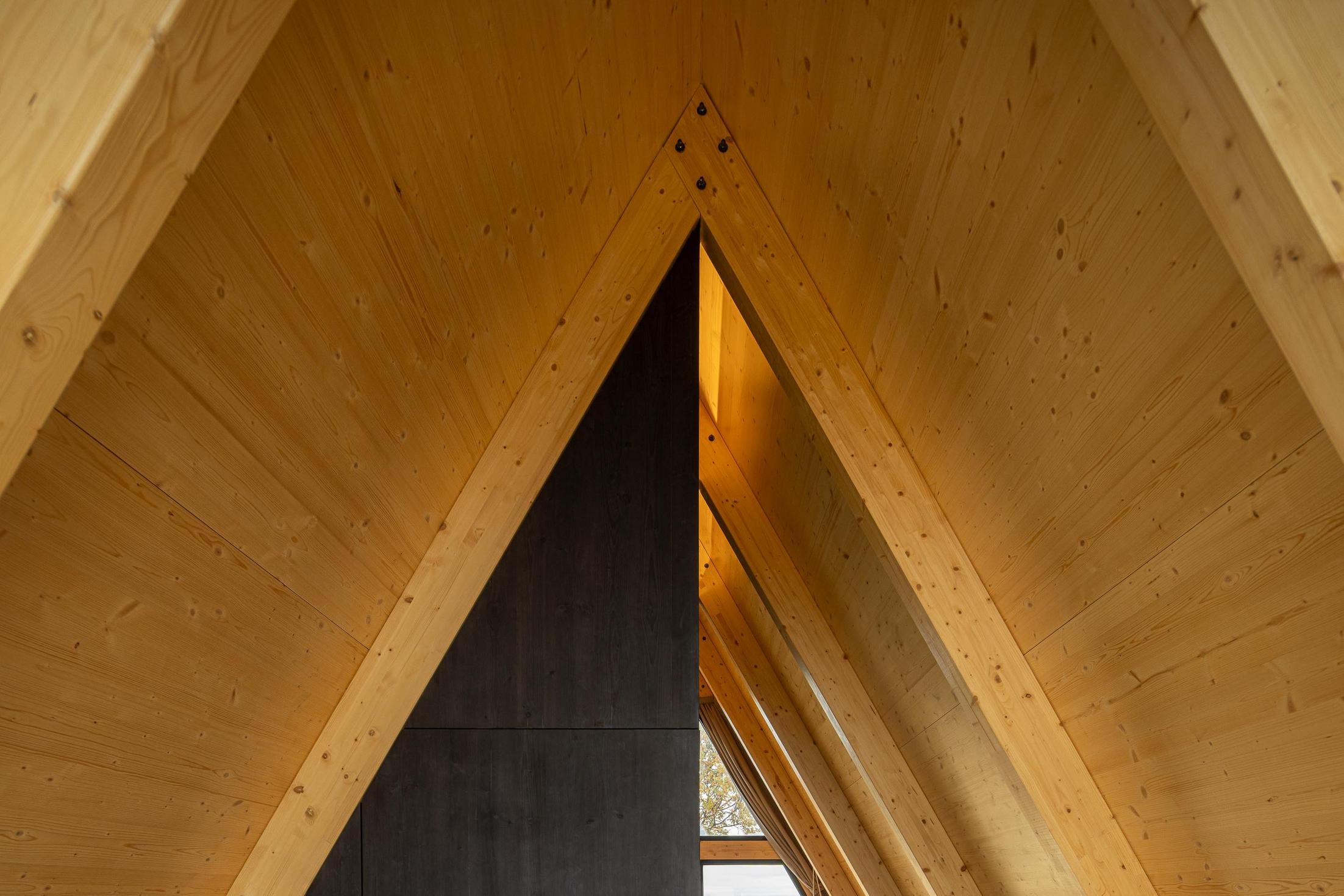 Interior com estrutura triangular de madeira clara no teto. Parede escura ao fundo. — Abrigos de Montanha arquitetura Filipe Pina Arquitectura em Belmonte