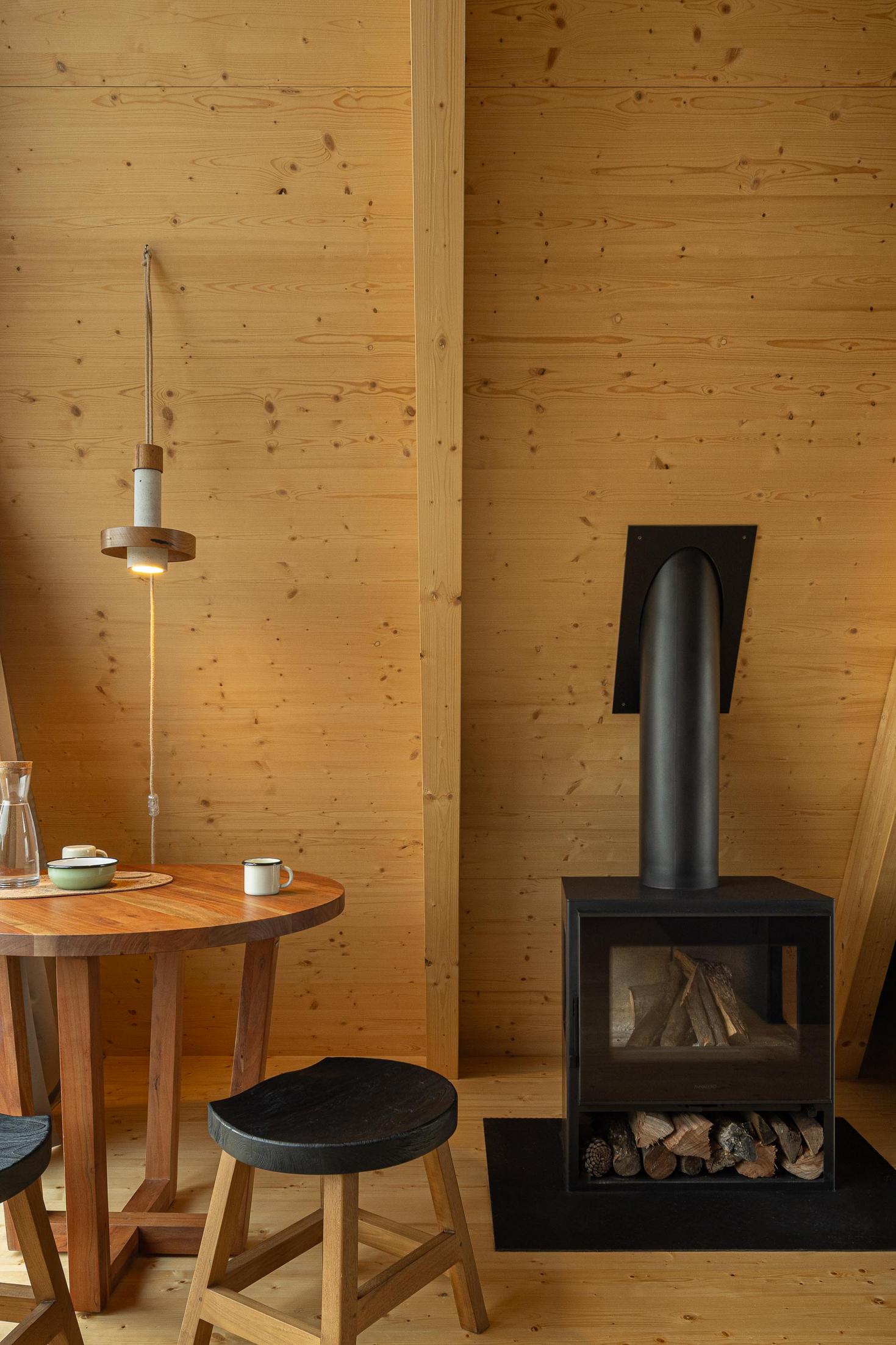 Sala de jantar com mesa redonda de madeira, cadeiras pretas, pavimento e paredes em madeira clara, e salamandra preta. Candeeiro suspenso. — Abrigos de Montanha arquitetura Filipe Pina Arquitectura em Belmonte