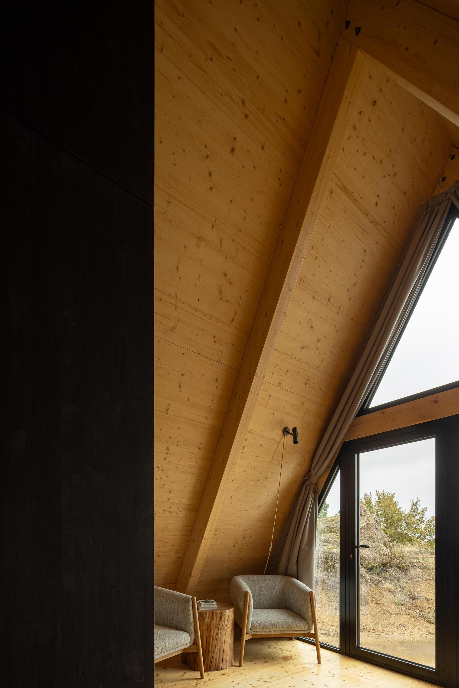 Zona de estar com cadeiras cinzentas, mesa de apoio em madeira, cortinas e pavimento de madeira clara. — Abrigos de Montanha arquitetura Filipe Pina Arquitectura em Belmonte