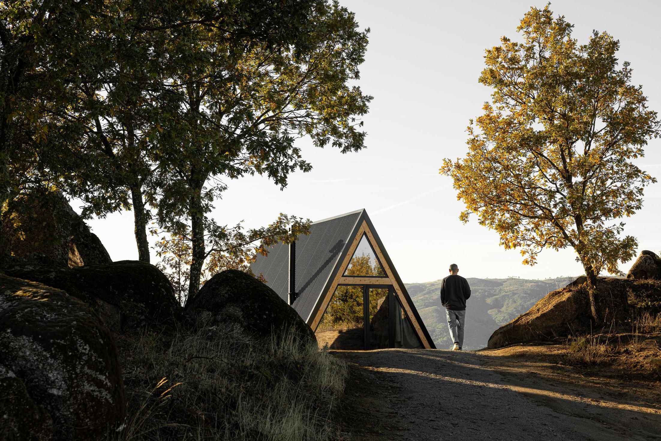 Exterior da cabana triangular, com homem a caminhar em direção à entrada. Paisagem rochosa e arborizada. — Abrigos de Montanha arquitetura Filipe Pina Arquitectura em Belmonte
