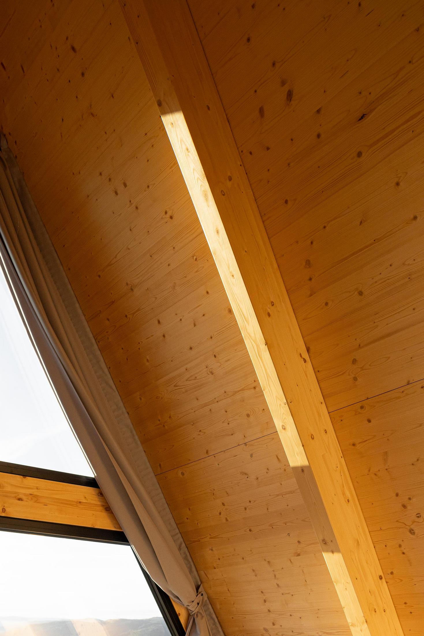 Detalhe do teto de madeira clara com cortina e viga. — Abrigos de Montanha arquitetura Filipe Pina Arquitectura em Belmonte
