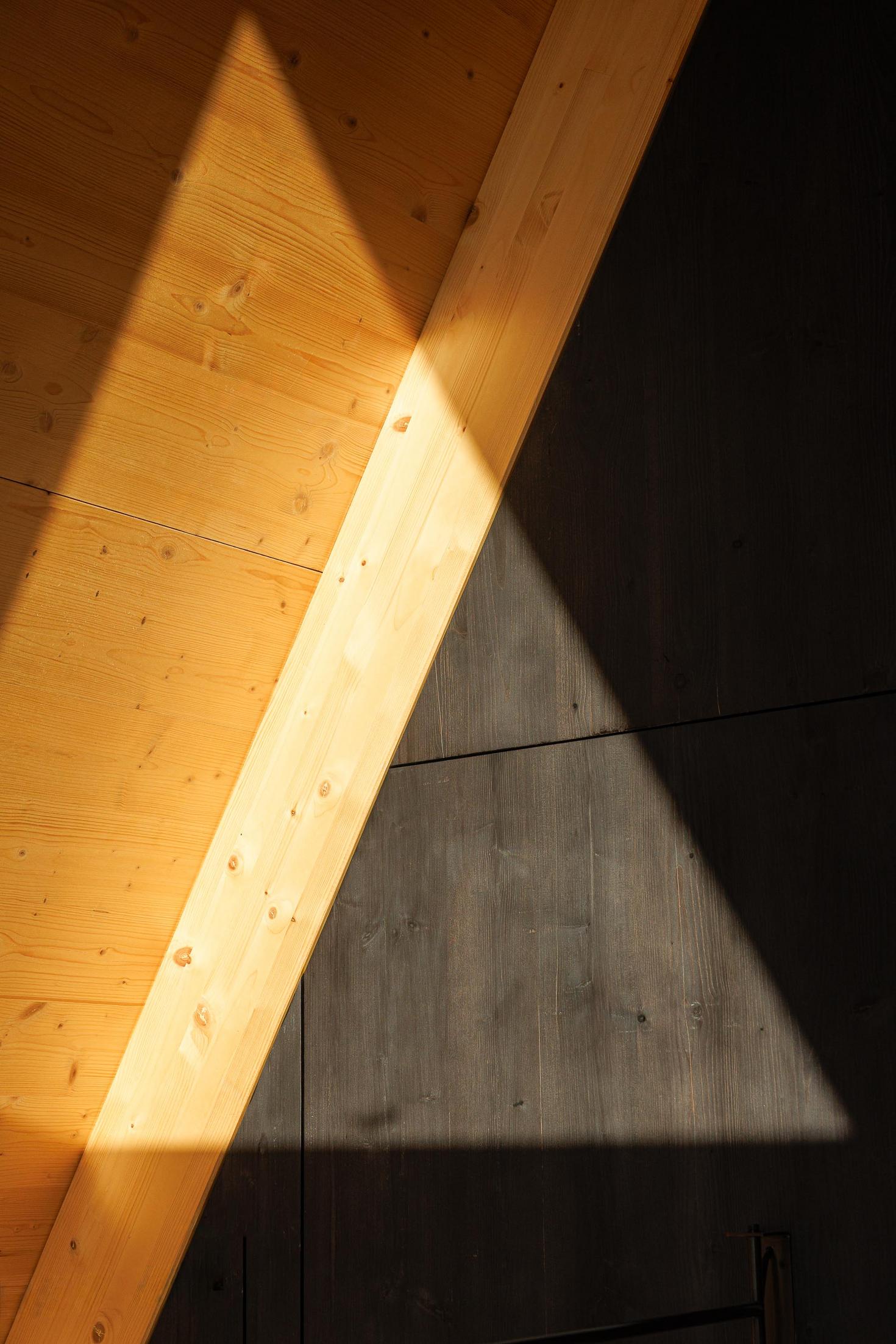 Detalhe da estrutura de madeira clara e parede escura, com luz solar a incidir. — Abrigos de Montanha arquitetura Filipe Pina Arquitectura em Belmonte