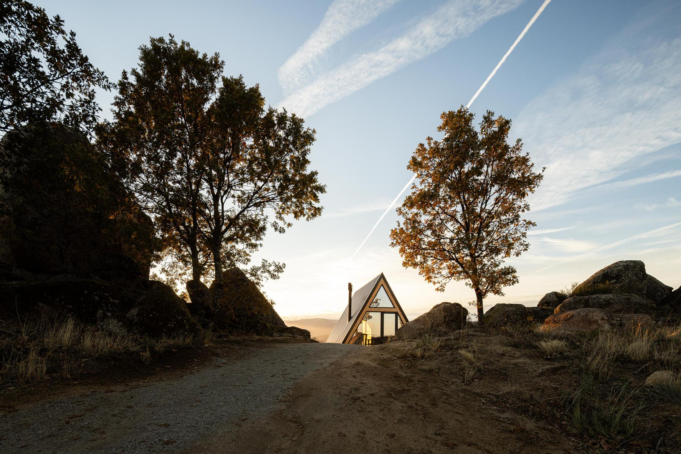Exterior da cabana triangular com árvores e rochas em primeiro plano. Céu azul com nuvens. — Abrigos de Montanha arquitetura Filipe Pina Arquitectura em Belmonte