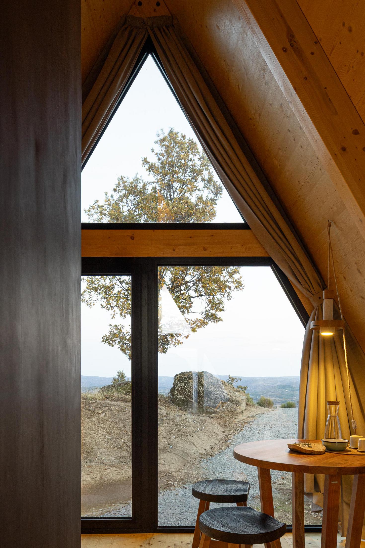 Sala de jantar com mesa redonda de madeira, candeeiro suspenso e vista para a paisagem exterior. — Abrigos de Montanha arquitetura Filipe Pina Arquitectura em Belmonte
