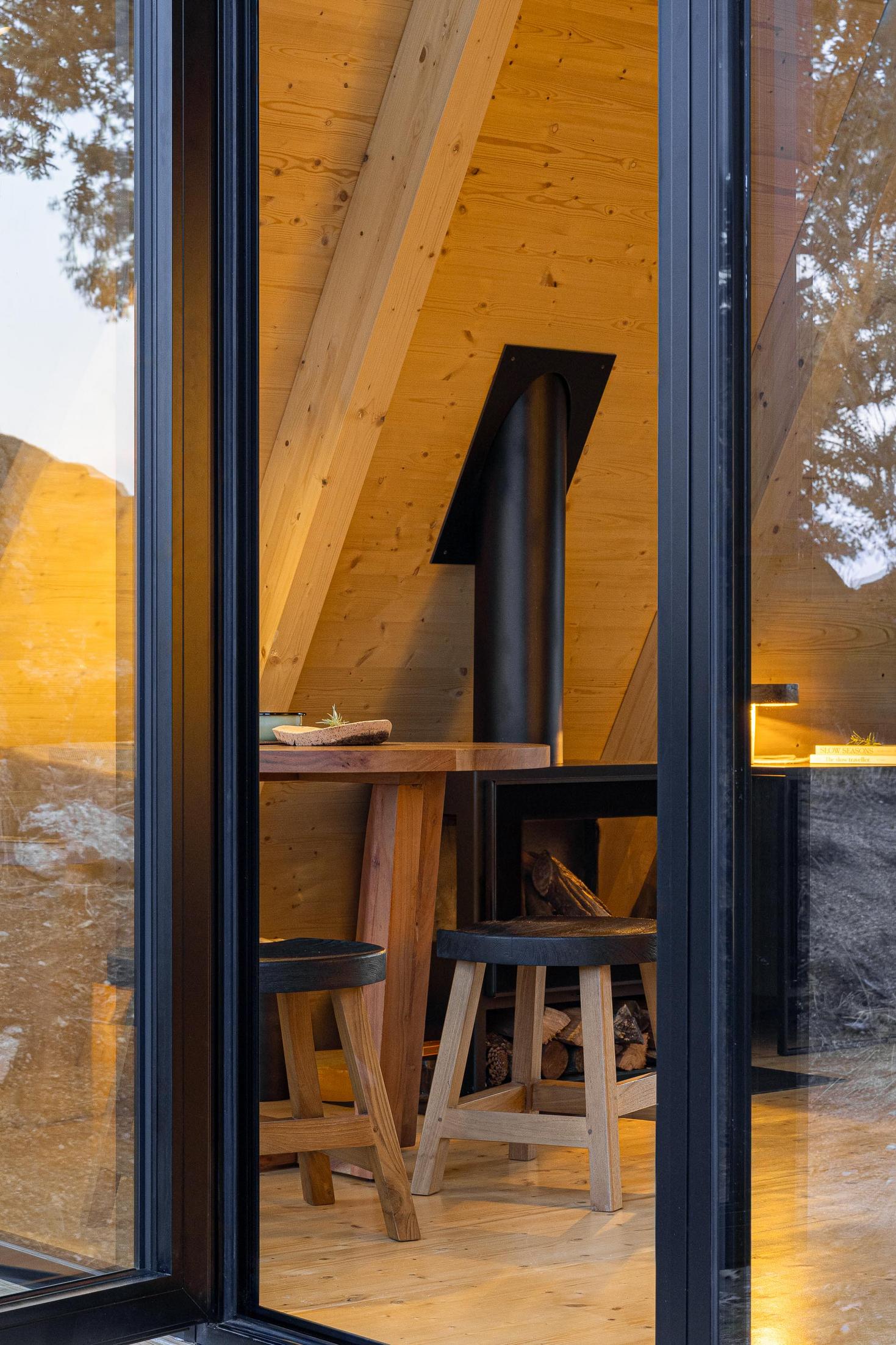 Sala de jantar com mesa redonda de madeira e salamandra preta, vista através da porta de vidro. — Abrigos de Montanha arquitetura Filipe Pina Arquitectura em Belmonte