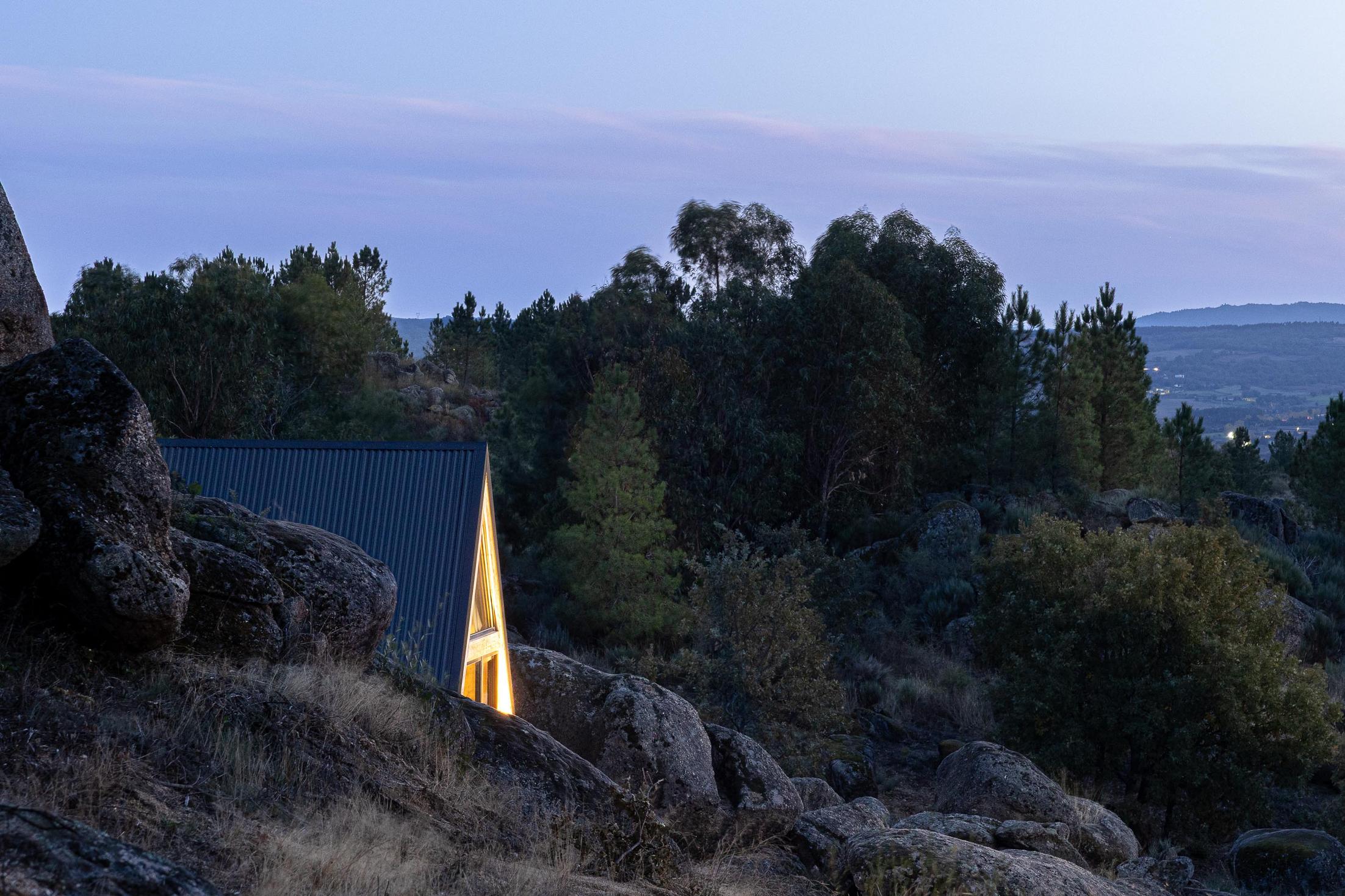 Exterior da cabana triangular com luz interior, vista entre rochas e vegetação ao anoitecer. — Abrigos de Montanha arquitetura Filipe Pina Arquitectura em Belmonte