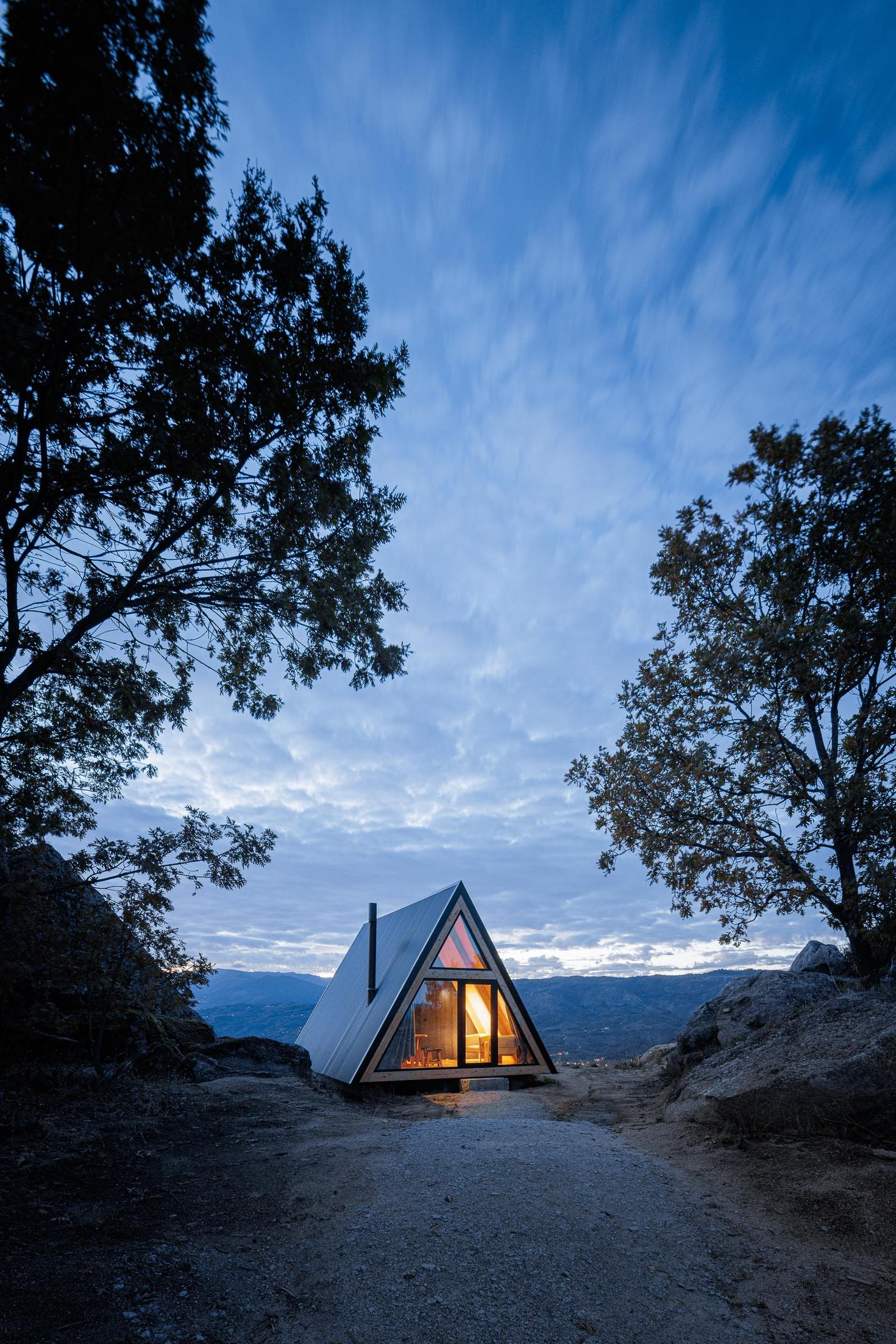 Exterior da cabana triangular iluminada à noite, rodeada por árvores e rochas. — Abrigos de Montanha arquitetura Filipe Pina Arquitectura em Belmonte
