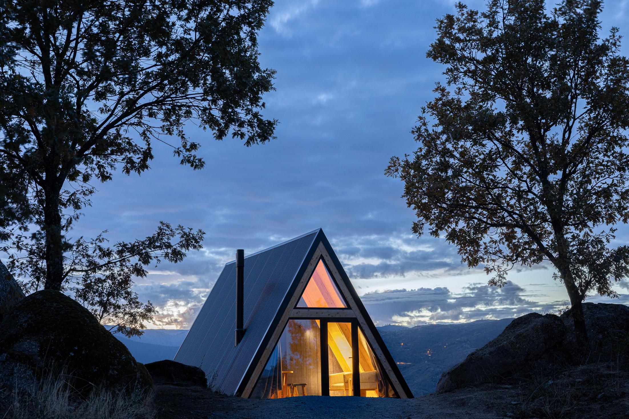 Exterior da cabana triangular iluminada à noite, com céu azul escuro e rochas em primeiro plano. — Abrigos de Montanha arquitetura Filipe Pina Arquitectura em Belmonte