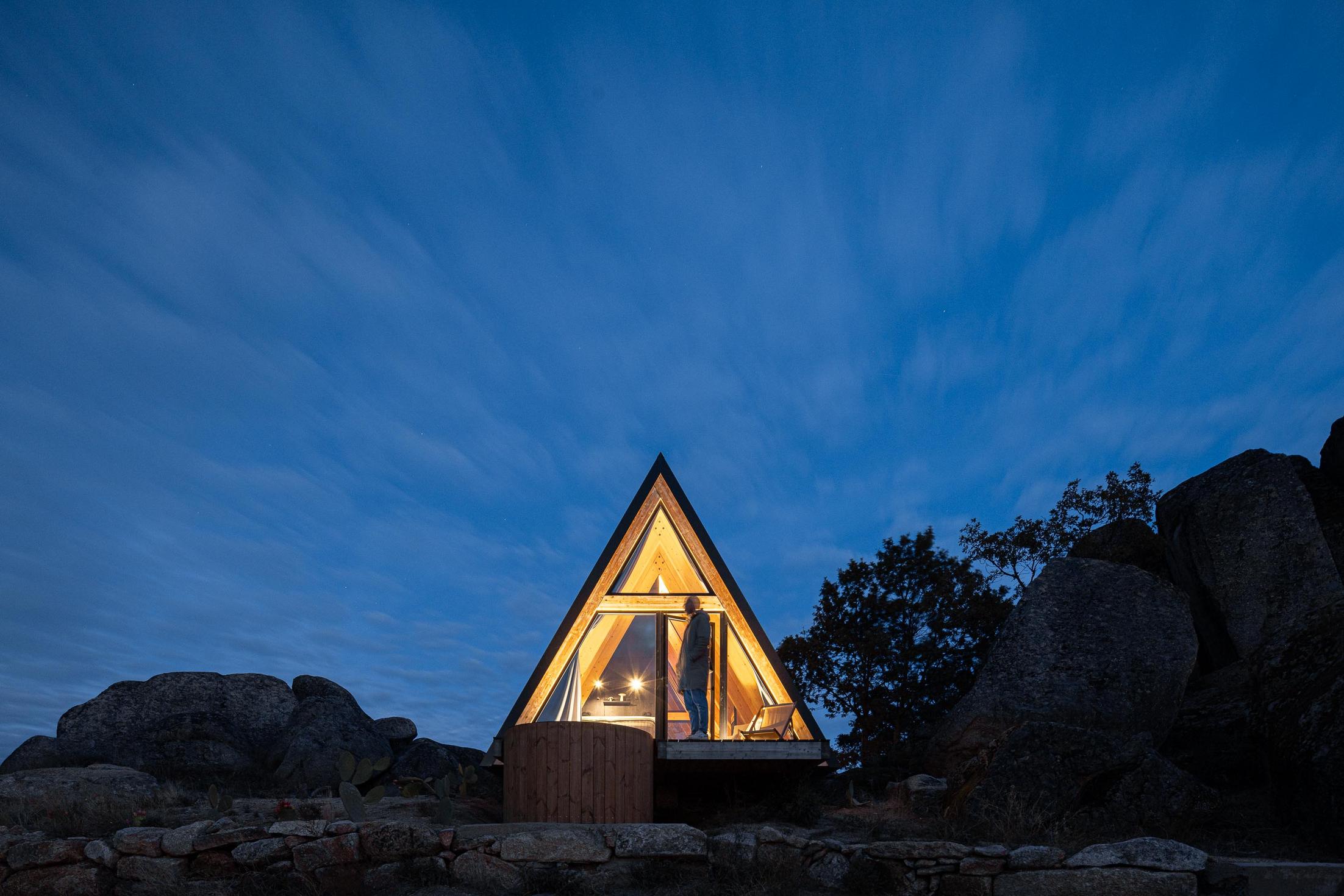 Exterior da cabana triangular iluminada à noite, com jacuzzi no deck. — Abrigos de Montanha arquitetura Filipe Pina Arquitectura em Belmonte