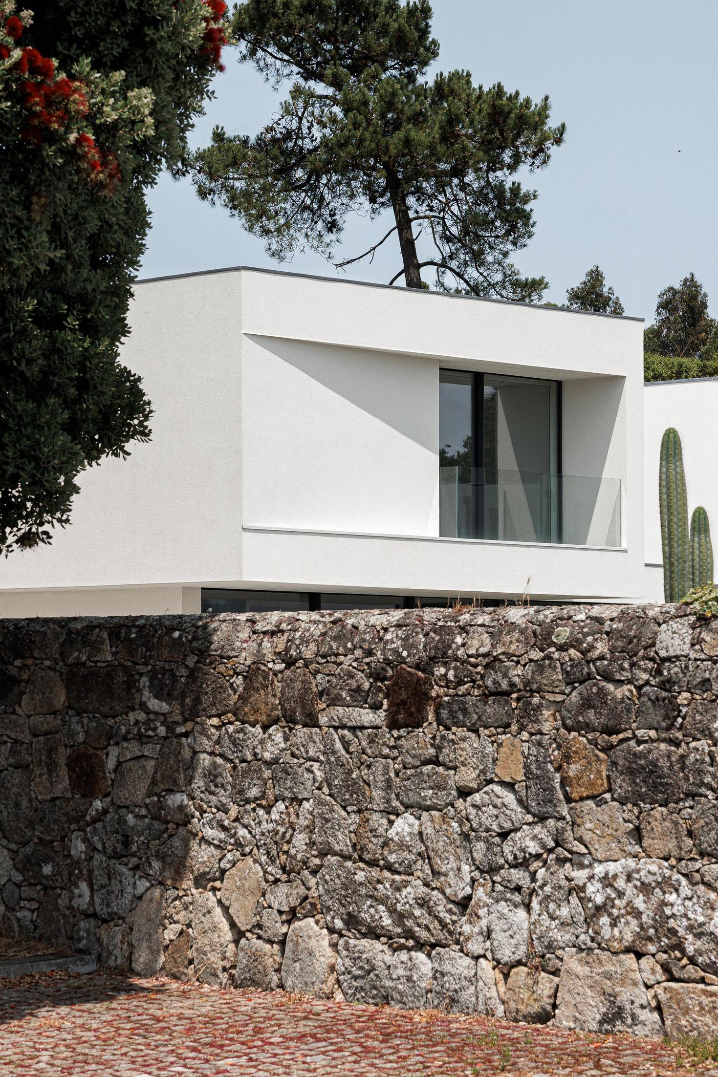 Fachada: Moradia minimalista branca com varanda envidraçada, sobre muro de pedra rústica e calçada. — Casa dos Sobreiros II arquitetura Urbanpolis em Póvoa de Varzim