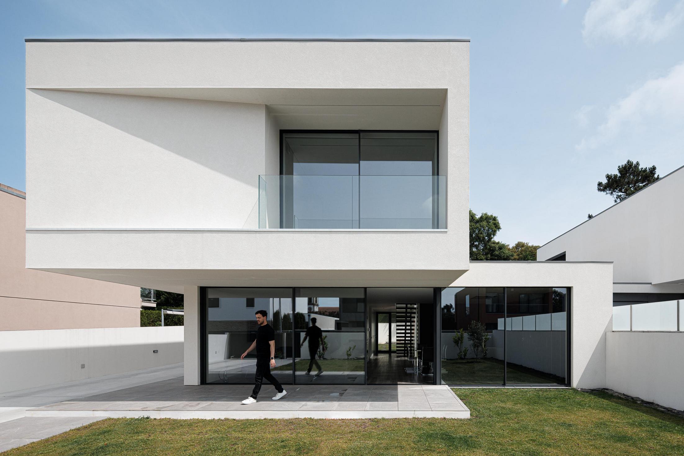 Exterior: Moradia branca de dois pisos com grandes janelas, relvado e homem a passear. — Casa dos Sobreiros II arquitetura Urbanpolis em Póvoa de Varzim
