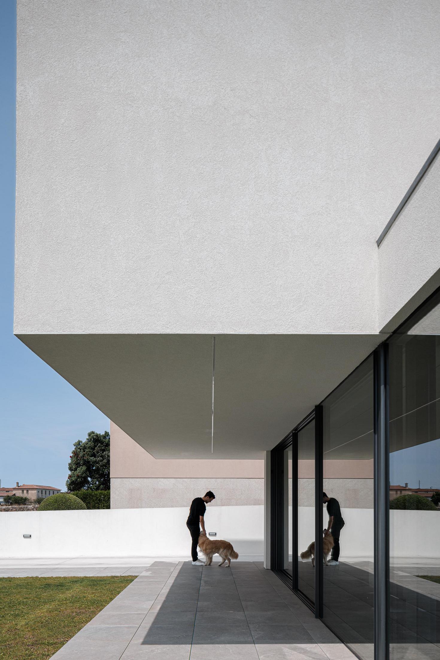 Exterior: Alpendre com pavimento cinzento, homem com cão e reflexos nas janelas. — Casa dos Sobreiros II arquitetura Urbanpolis em Póvoa de Varzim