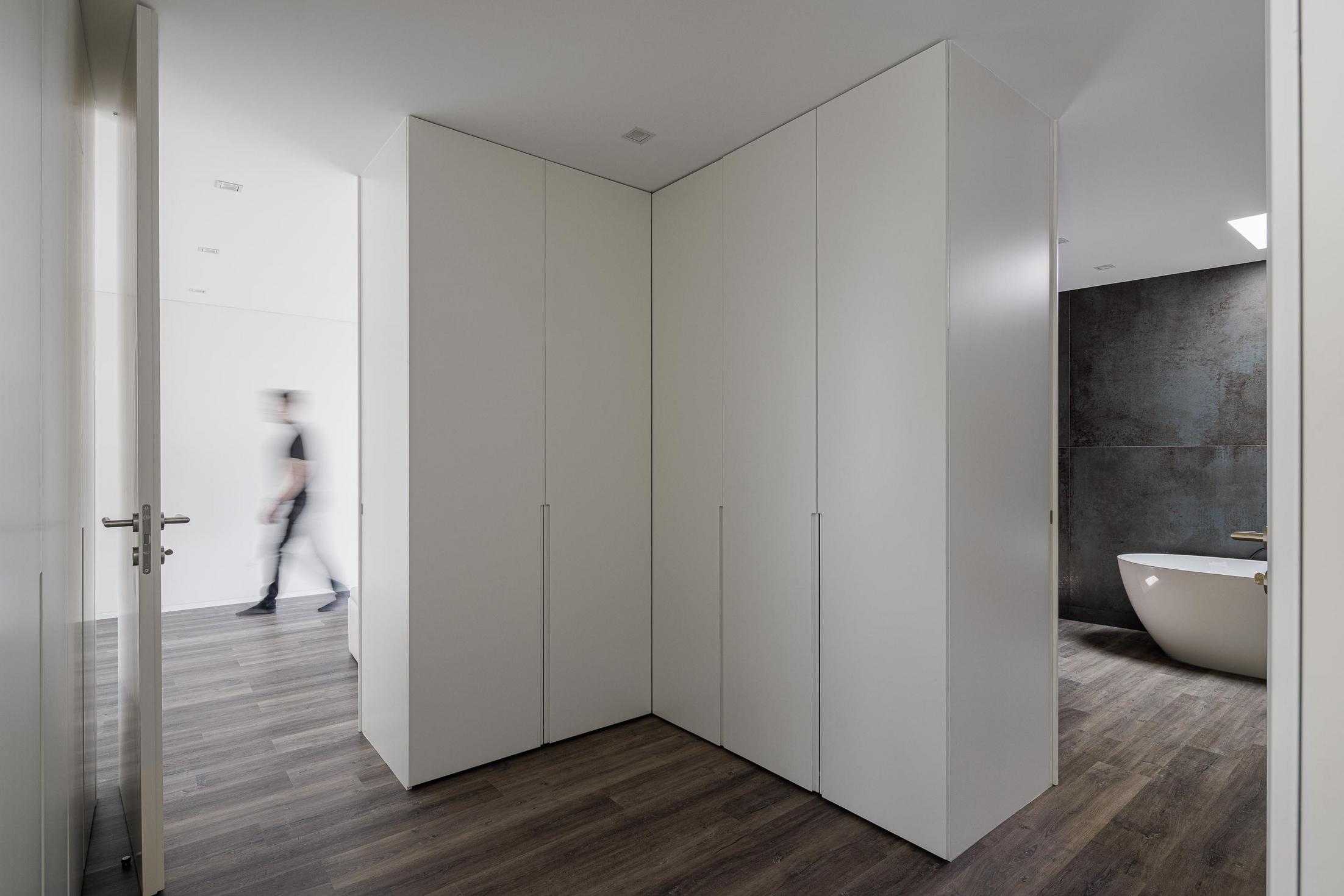 Closet/Casa de banho: Roupeiro branco, corredor e banheira branca numa casa de banho com luz zenital. — Casa dos Sobreiros II arquitetura Urbanpolis em Póvoa de Varzim