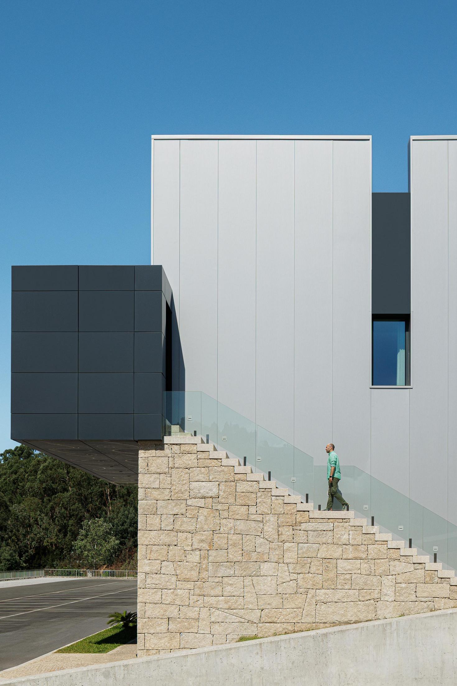 Escadaria exterior: Degraus em pedra rústica com guarda em vidro, acesso a volume saliente revestido a painéis escuros. — Bradco II arquitetura Em Paralelo em Castelo de Paiva