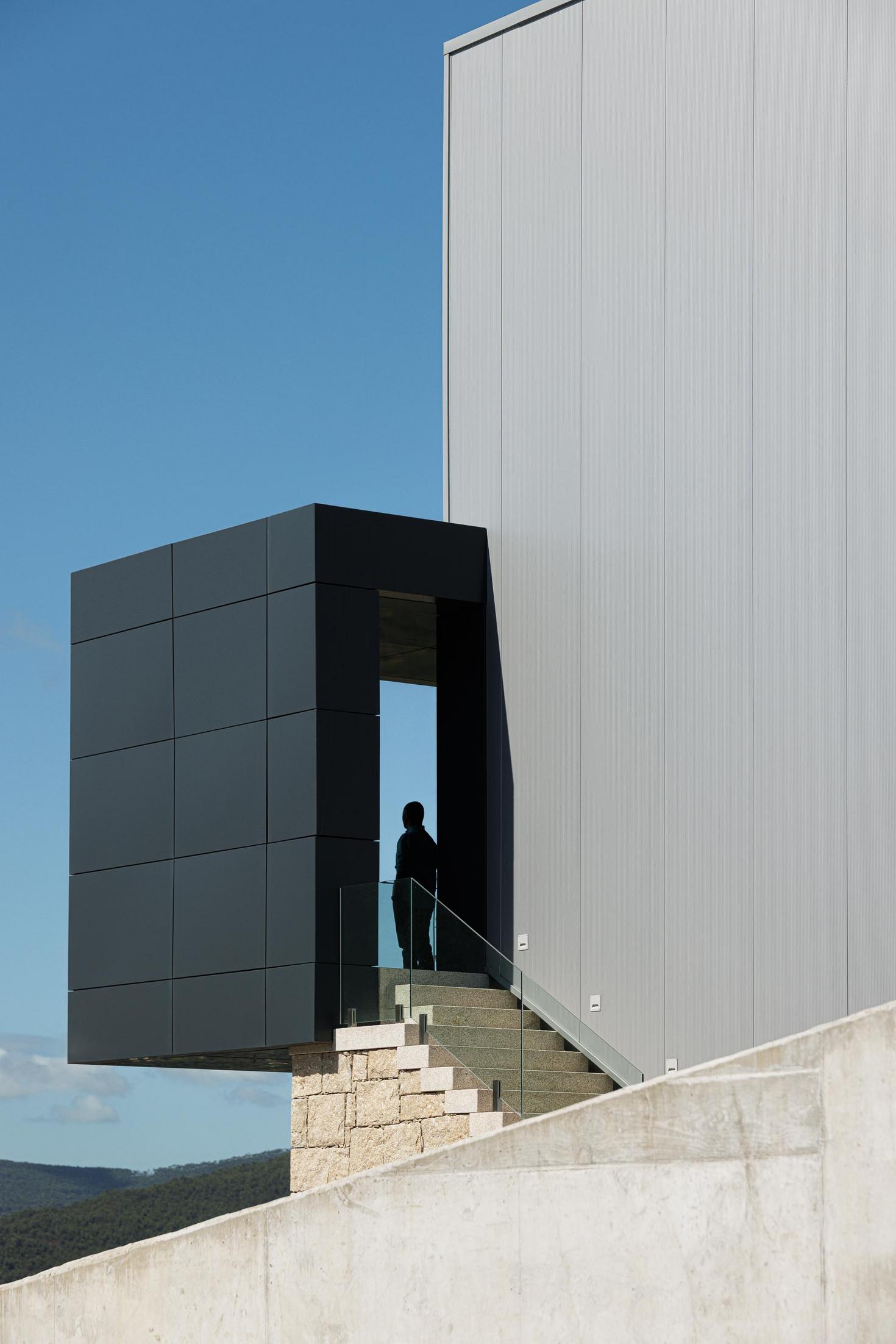 Escadaria exterior: Vista inferior do volume saliente, com pessoa na entrada, e escadaria em pedra. — Bradco II arquitetura Em Paralelo em Castelo de Paiva