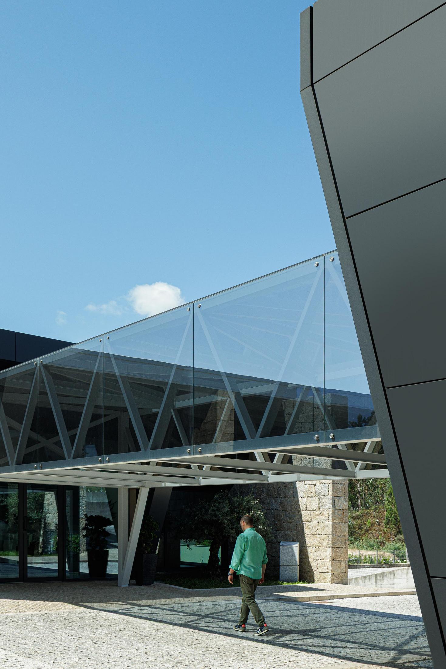 Passarela: Estrutura envidraçada que liga dois edifícios com revestimento em pedra e metal. — Bradco II arquitetura Em Paralelo em Castelo de Paiva