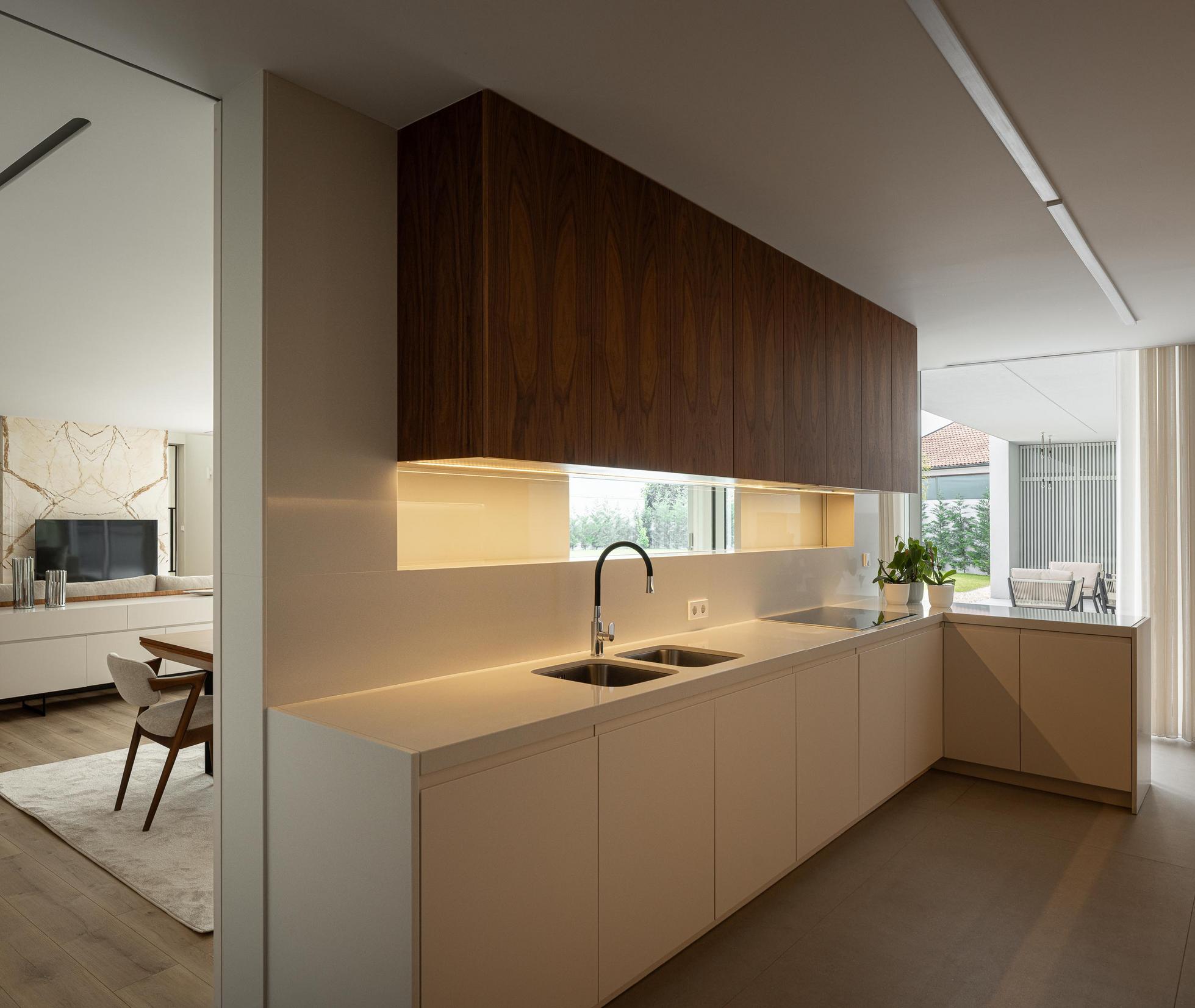 Cozinha: Armários superiores em madeira escura, bancada branca e vista para a sala de estar. — Casa Branca Minimalista arquitetura Arquitetura501 em Braga