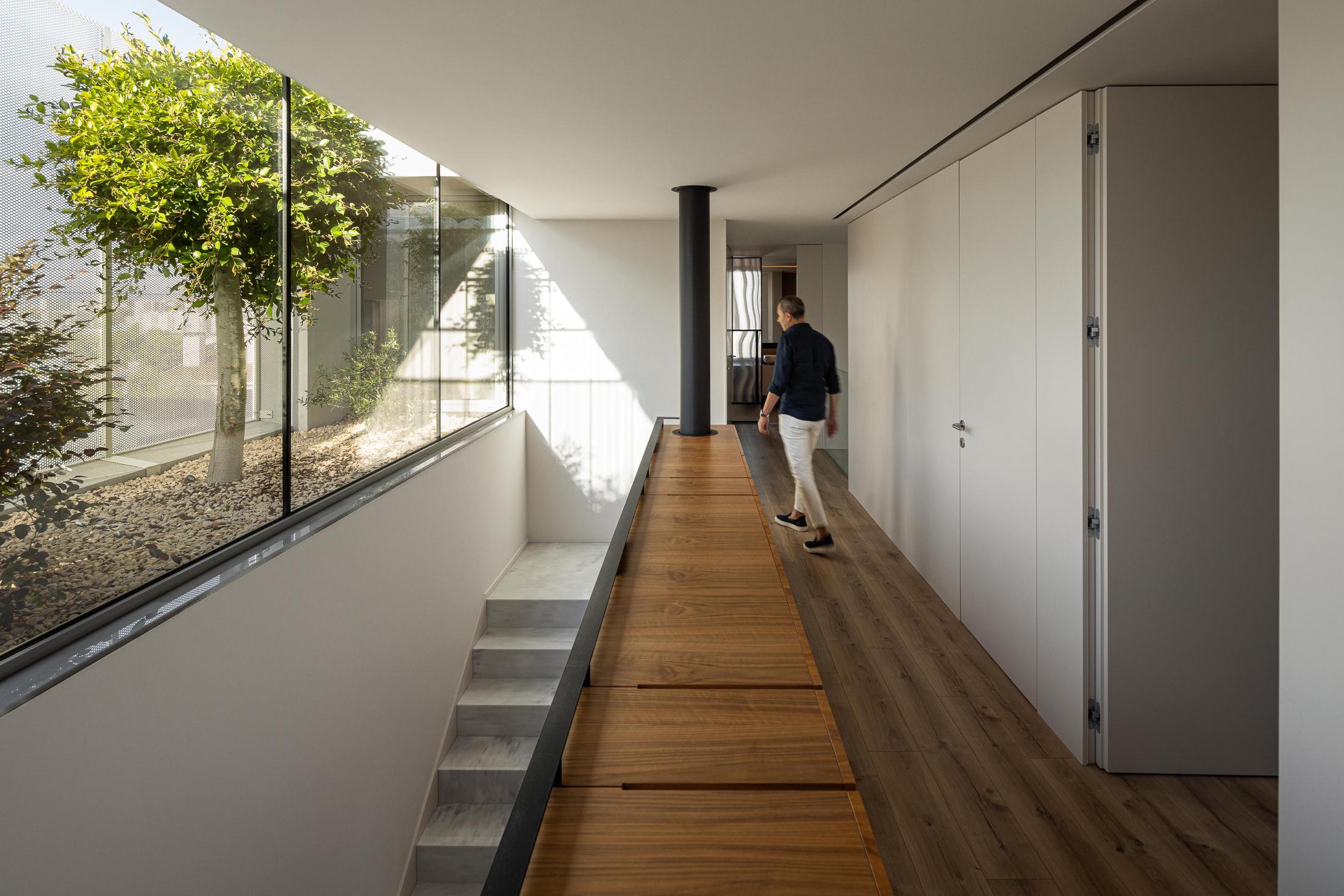 Corredor superior: Pessoa a caminhar no corredor com vista para o jardim interior e escadas. — Casa Branca Minimalista arquitetura Arquitetura501 em Braga