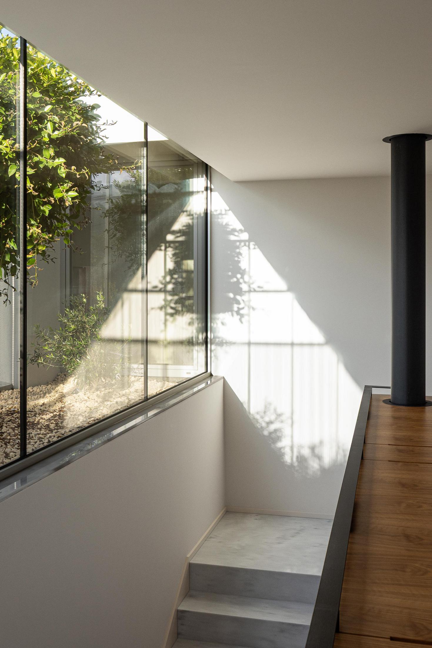 Corredor superior: Janela envidraçada com vista para jardim interior e escadas em mármore. — Casa Branca Minimalista arquitetura Arquitetura501 em Braga