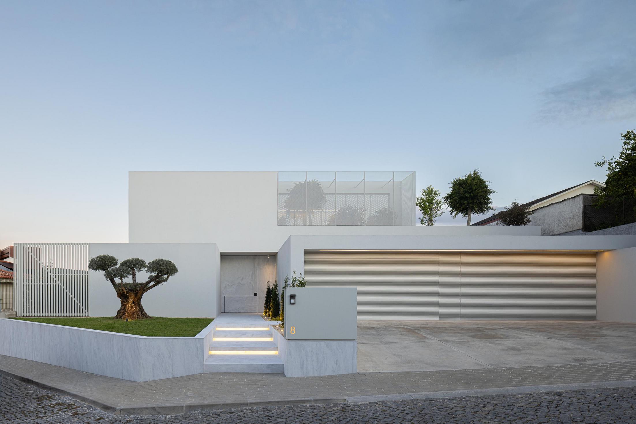 Exterior: Vista frontal da casa com garagem, entrada principal e jardim com oliveira. — Casa Branca Minimalista arquitetura Arquitetura501 em Braga