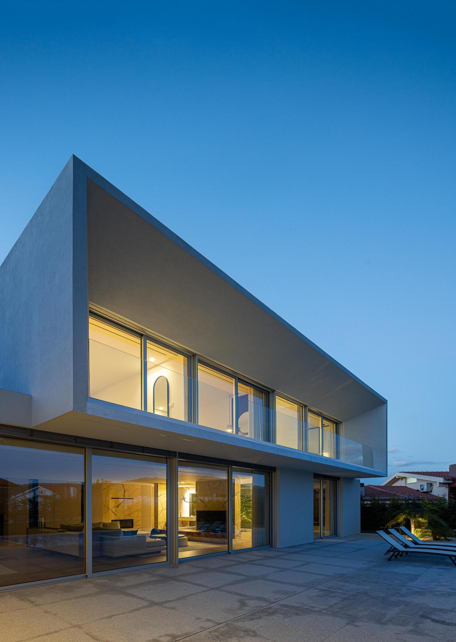 Exterior: Fachada posterior da casa ao entardecer, com iluminação interior e exterior. — Casa Branca Minimalista arquitetura Arquitetura501 em Braga