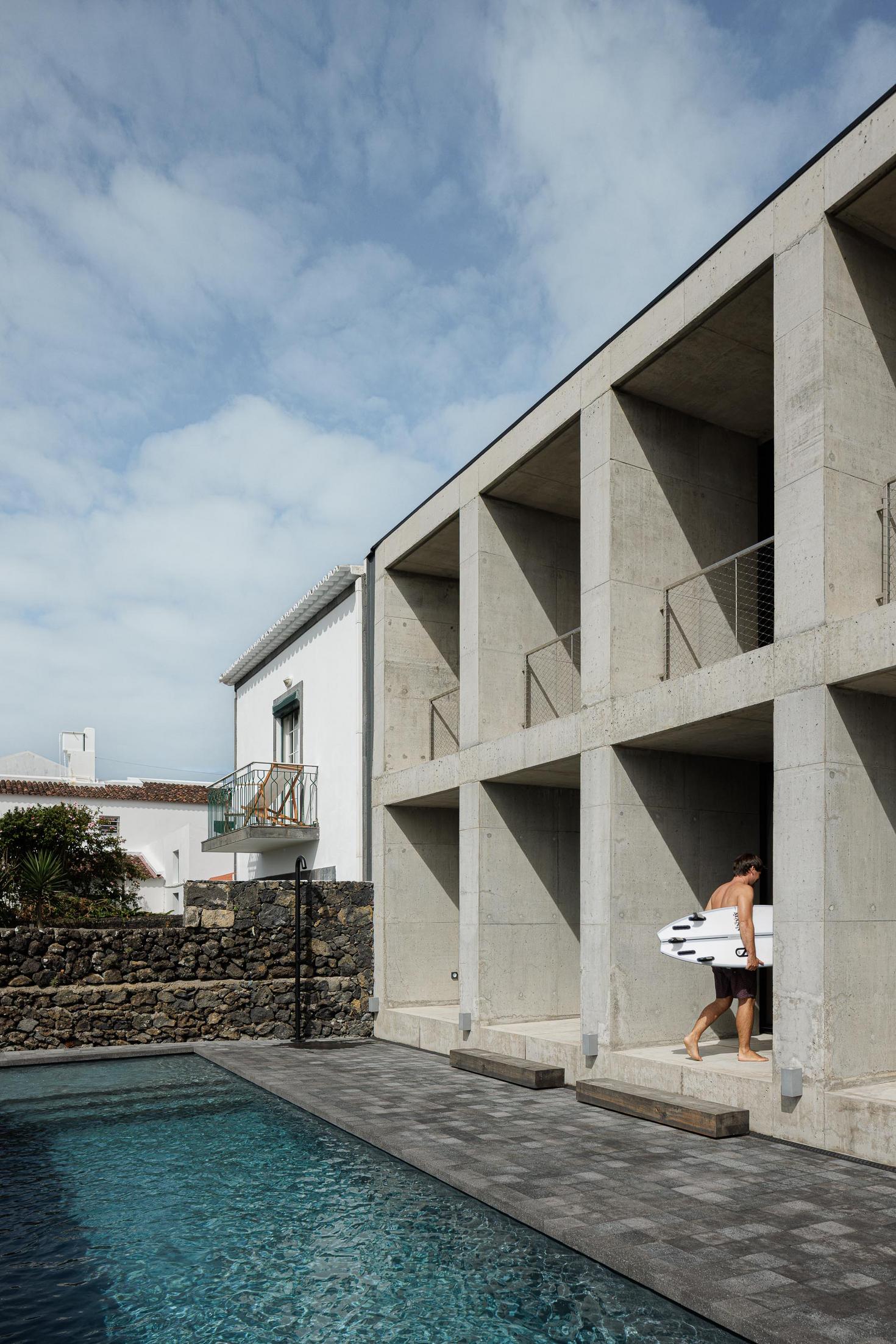 Fachada: Edifício em betão com piscina. Homem com prancha de surf entra na casa. — Casa da Rocha Quebrada arquitetura SO Arquitetura & Design em São Miguel