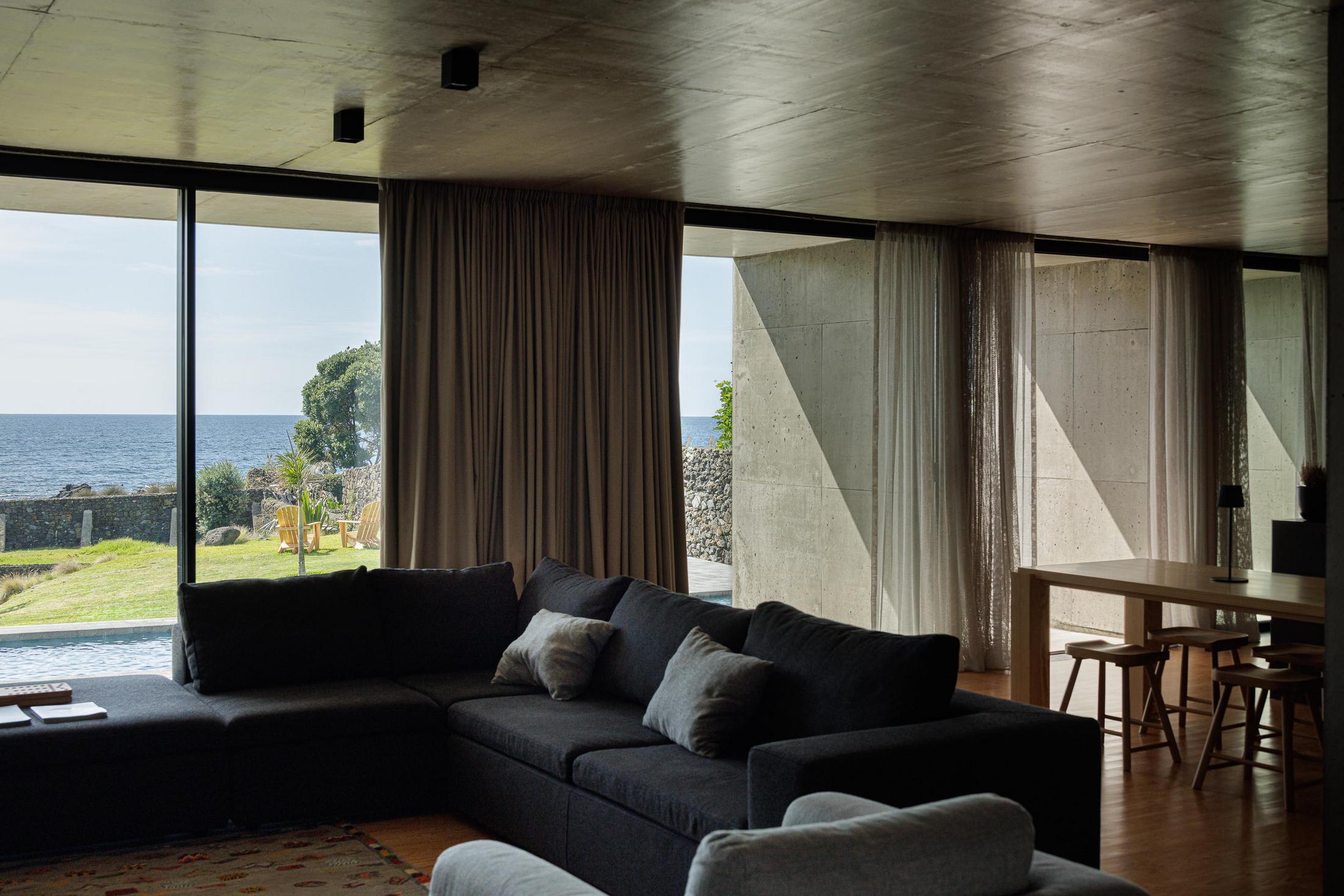 Sala de estar: Sofá cinzento e mesa de jantar em madeira. Vista para o jardim e mar. — Casa da Rocha Quebrada arquitetura SO Arquitetura & Design em São Miguel