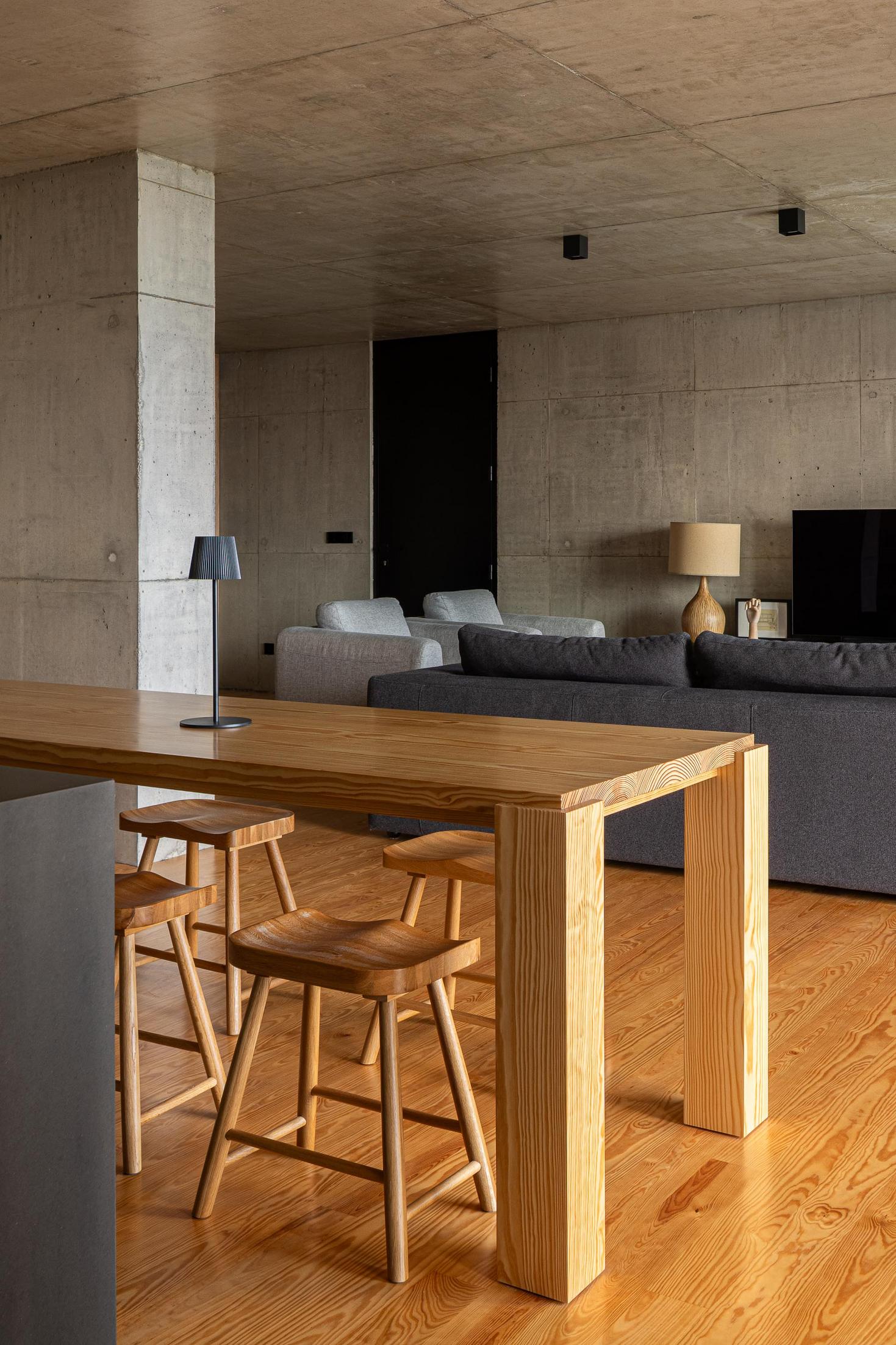 Sala de estar: Mesa de madeira com bancos e candeeiro. Sofá cinzento e coluna de betão. — Casa da Rocha Quebrada arquitetura SO Arquitetura & Design em São Miguel