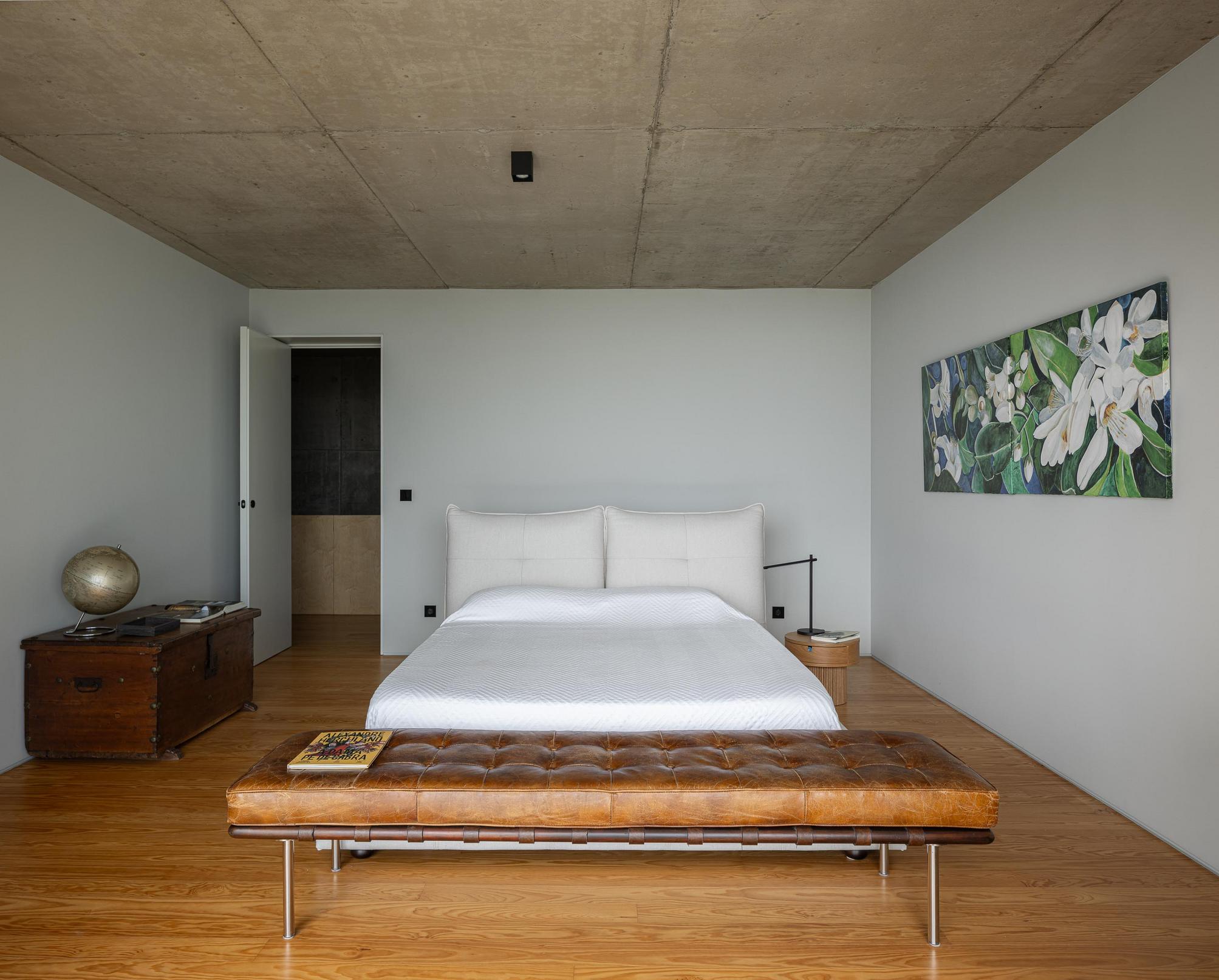 Quarto: Cama branca com cabeceira almofadada. Pavimento em madeira e parede cinzenta com pintura. — Casa da Rocha Quebrada arquitetura SO Arquitetura & Design em São Miguel