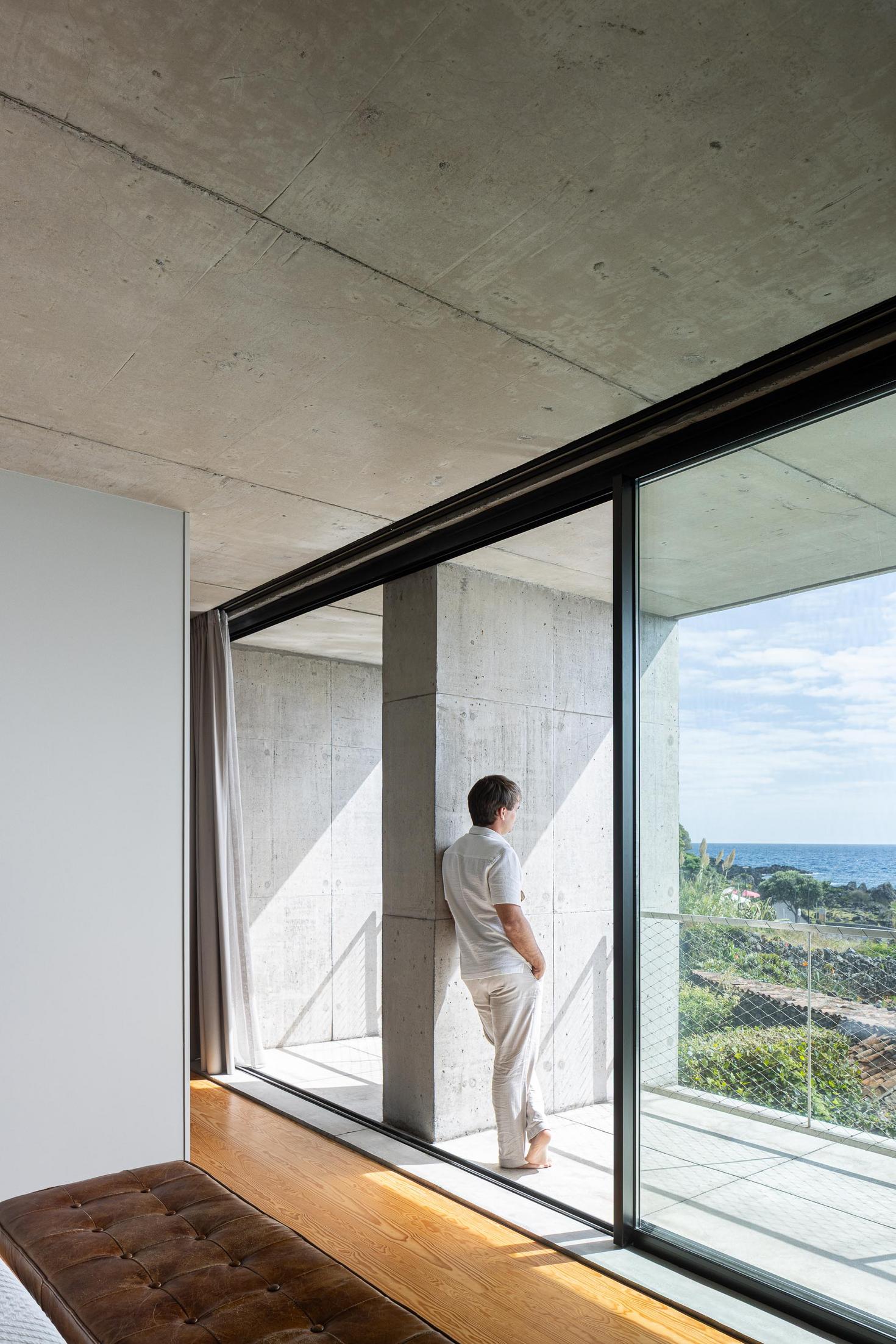 Quarto: Homem olha para a varanda. Cama branca, banco em pele e pavimento em madeira. — Casa da Rocha Quebrada arquitetura SO Arquitetura & Design em São Miguel