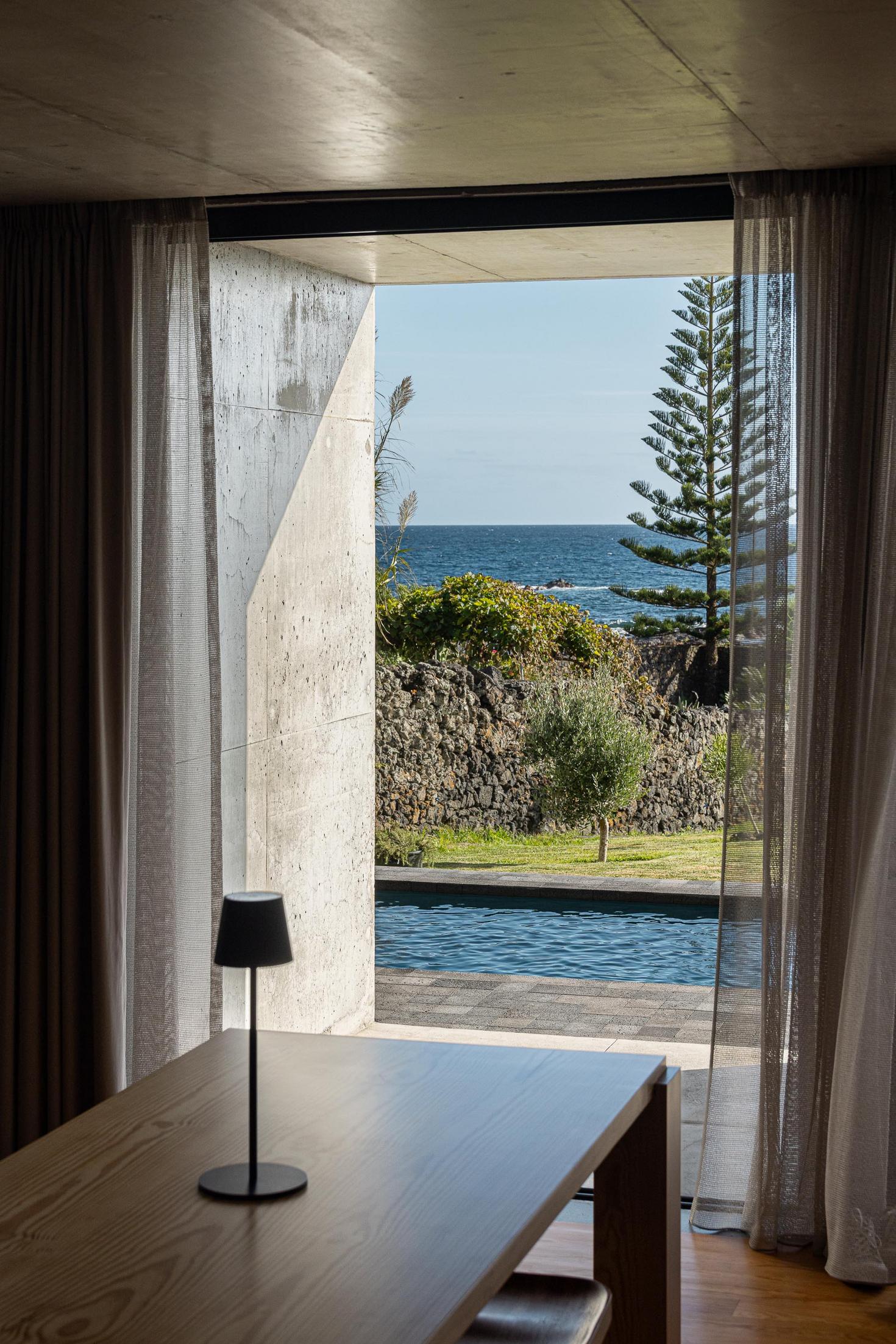 Sala de jantar: Mesa em madeira com vista para o mar. Cortinas claras e candeeiro preto. — Casa da Rocha Quebrada arquitetura SO Arquitetura & Design em São Miguel