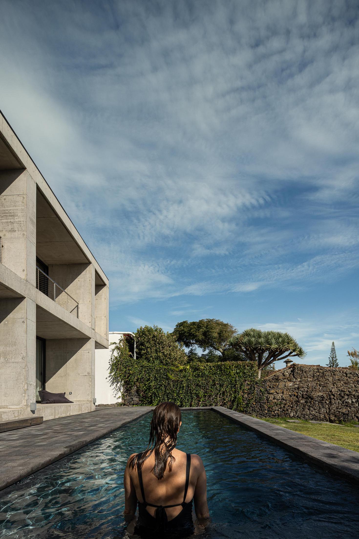 Exterior: Mulher na piscina com vista para o edifício em betão. Vegetação e céu azul. — Casa da Rocha Quebrada arquitetura SO Arquitetura & Design em São Miguel