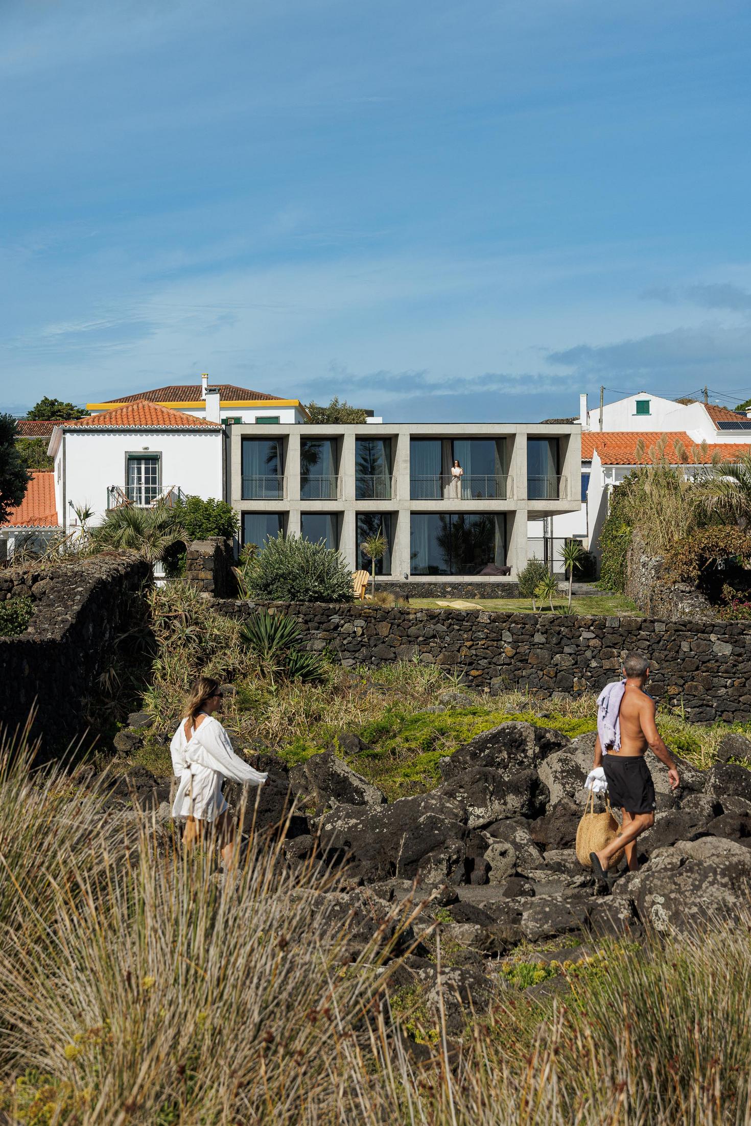 Exterior: Edifício em betão visto das rochas. Vegetação rasteira e casas tradicionais ao fundo. — Casa da Rocha Quebrada arquitetura SO Arquitetura & Design em São Miguel