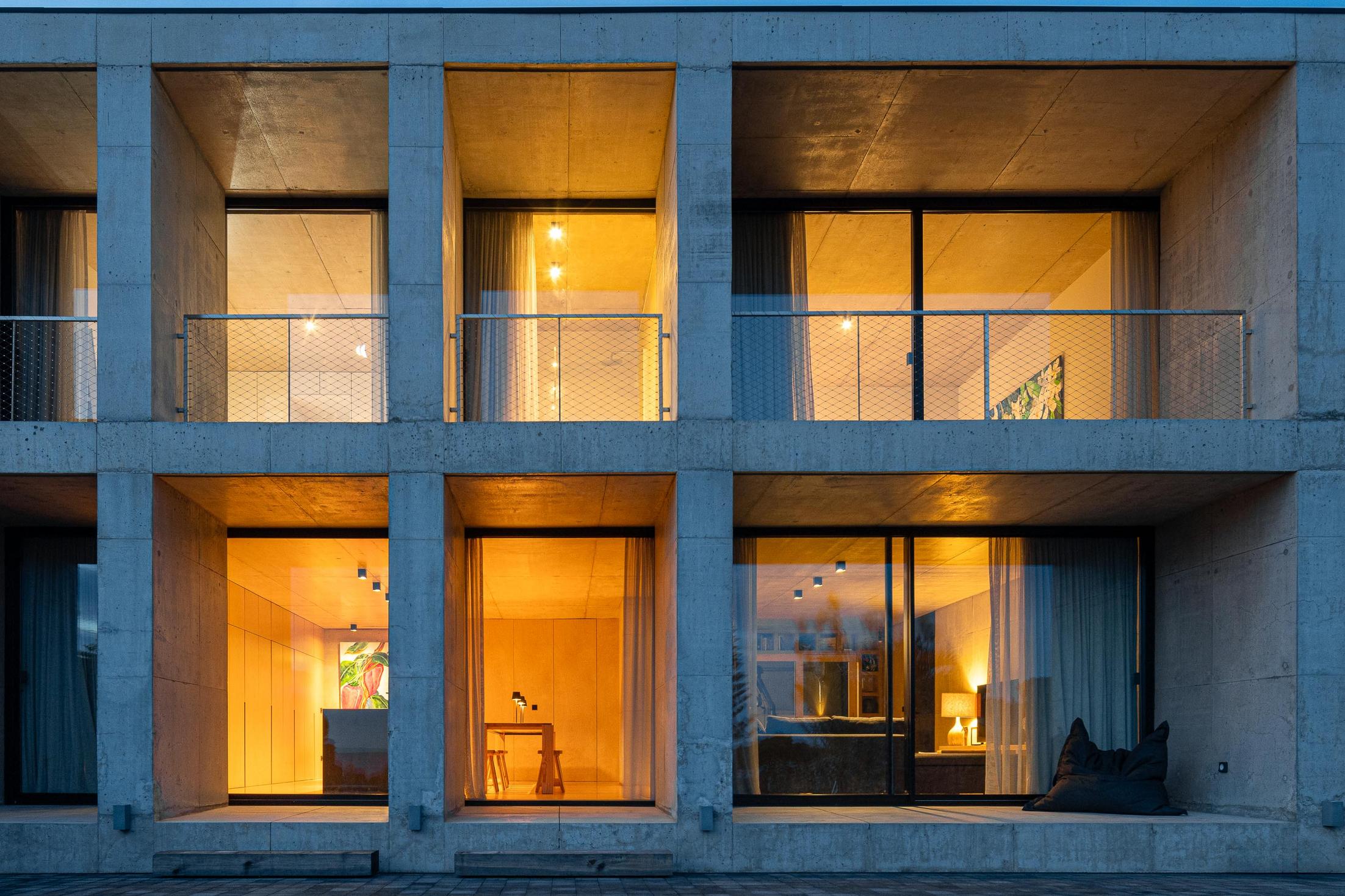 Fachada: Edifício em betão com janelas iluminadas. Interior moderno com madeira e candeeiros. — Casa da Rocha Quebrada arquitetura SO Arquitetura & Design em São Miguel