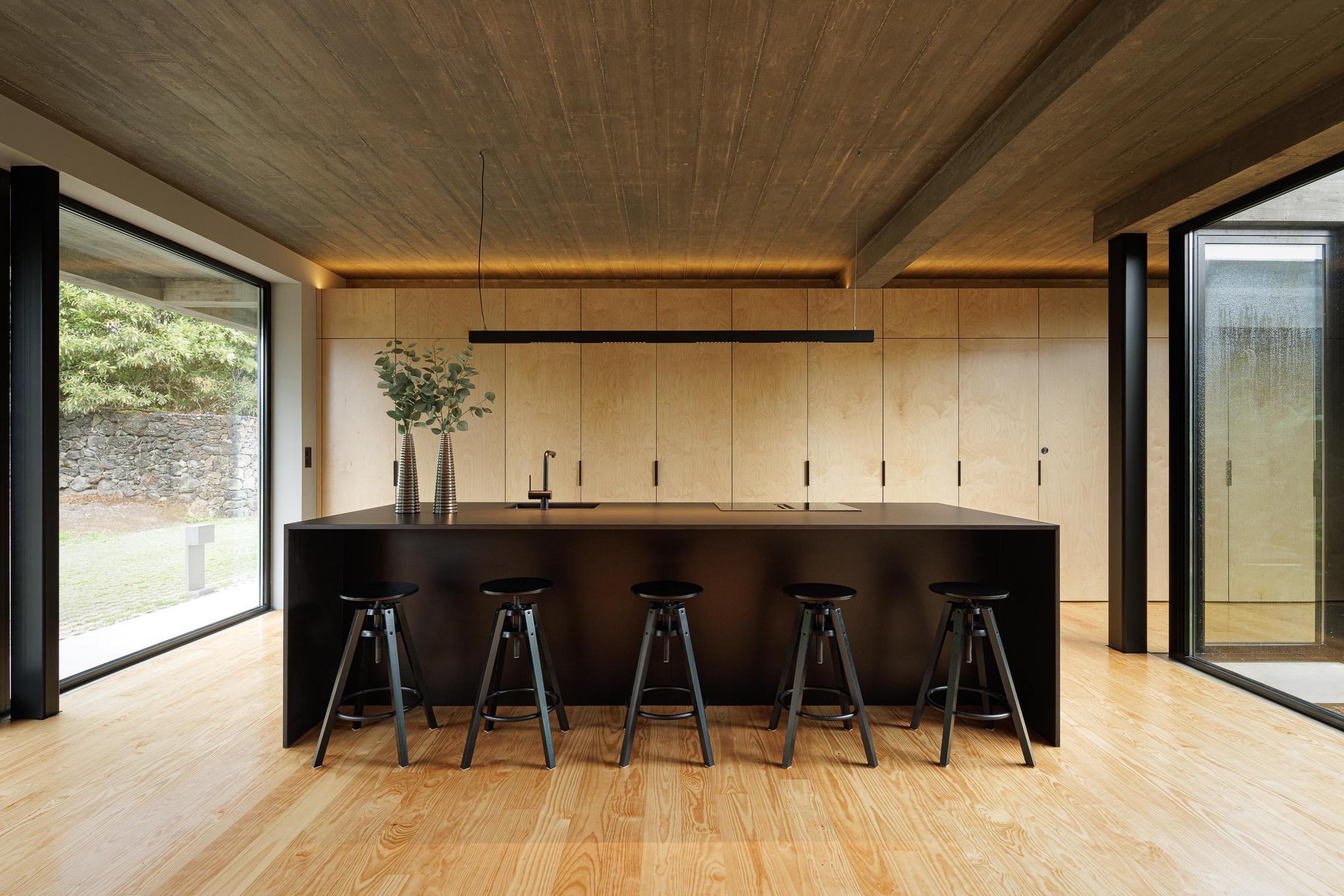 Cozinha: Ilha central preta com bancos, armários em contraplacado e pavimento em madeira clara. Candeeiro suspenso linear. — Casa CR arquitetura SO Arquitetura & Design em São Miguel