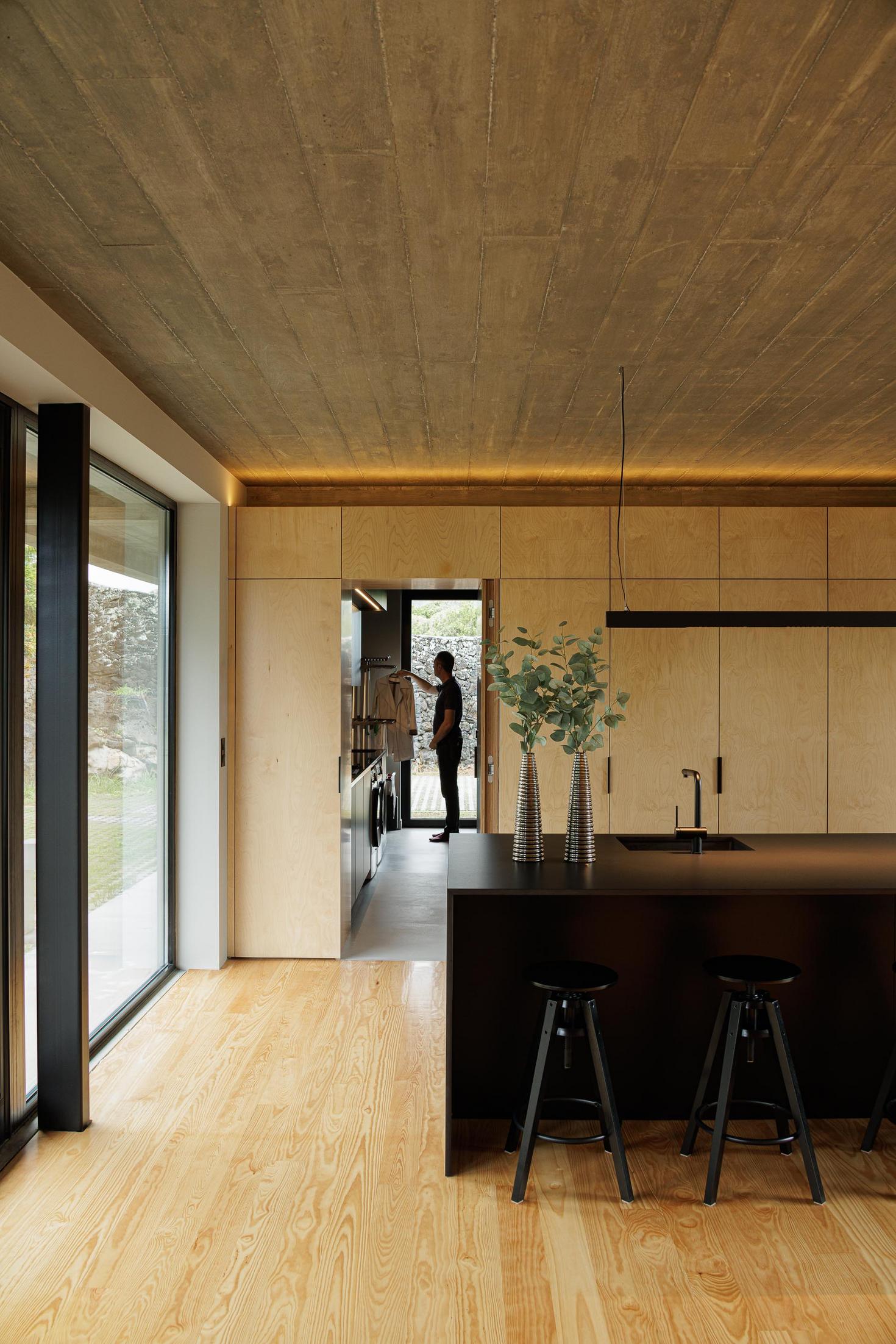 Cozinha: Ilha central preta, armários em contraplacado e pavimento em madeira. Pessoa a entrar na cozinha. — Casa CR arquitetura SO Arquitetura & Design em São Miguel